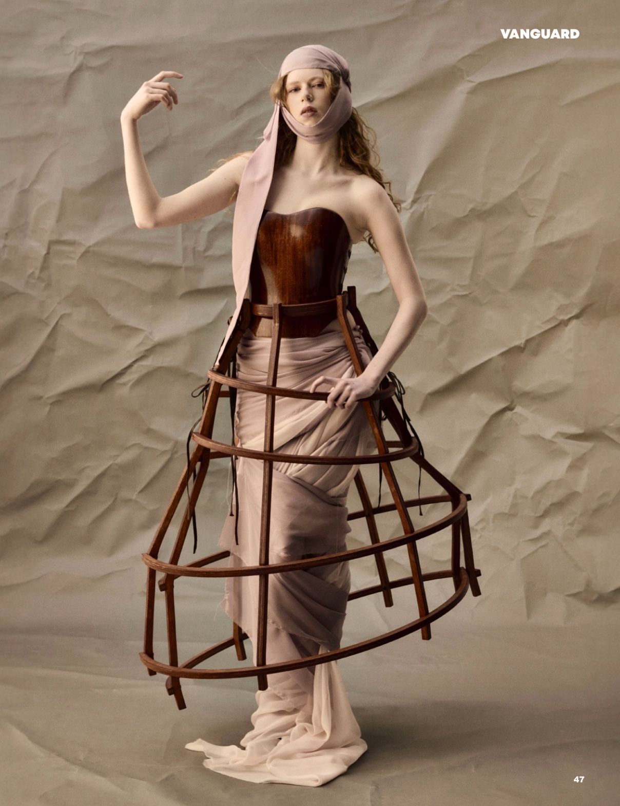 A woman in a period-style dress with a cage-like structure underneath stands against a backdrop of crumpled paper. Her hand is raised in a dramatic gesture.