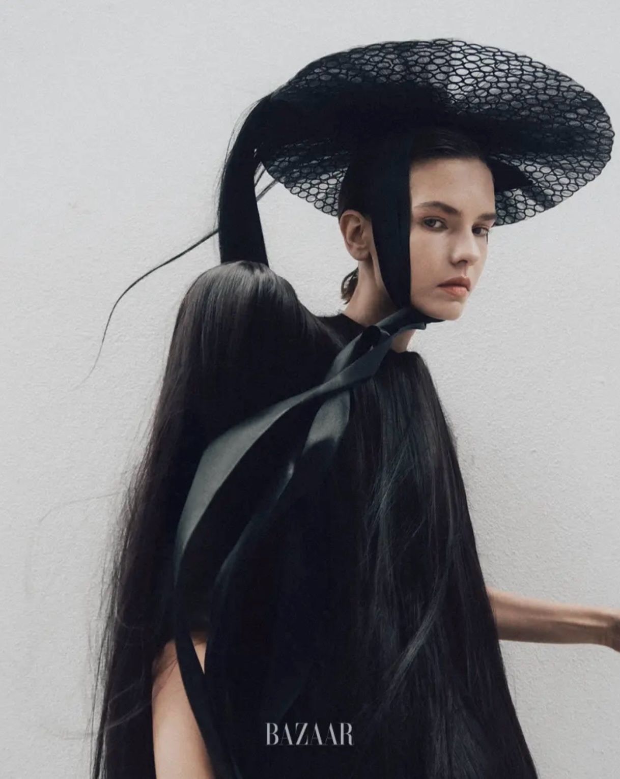 A woman wearing an extravagant black hat and a long, flowing dress against a white background. The image features a BAZAAR logo in the bottom right corner.