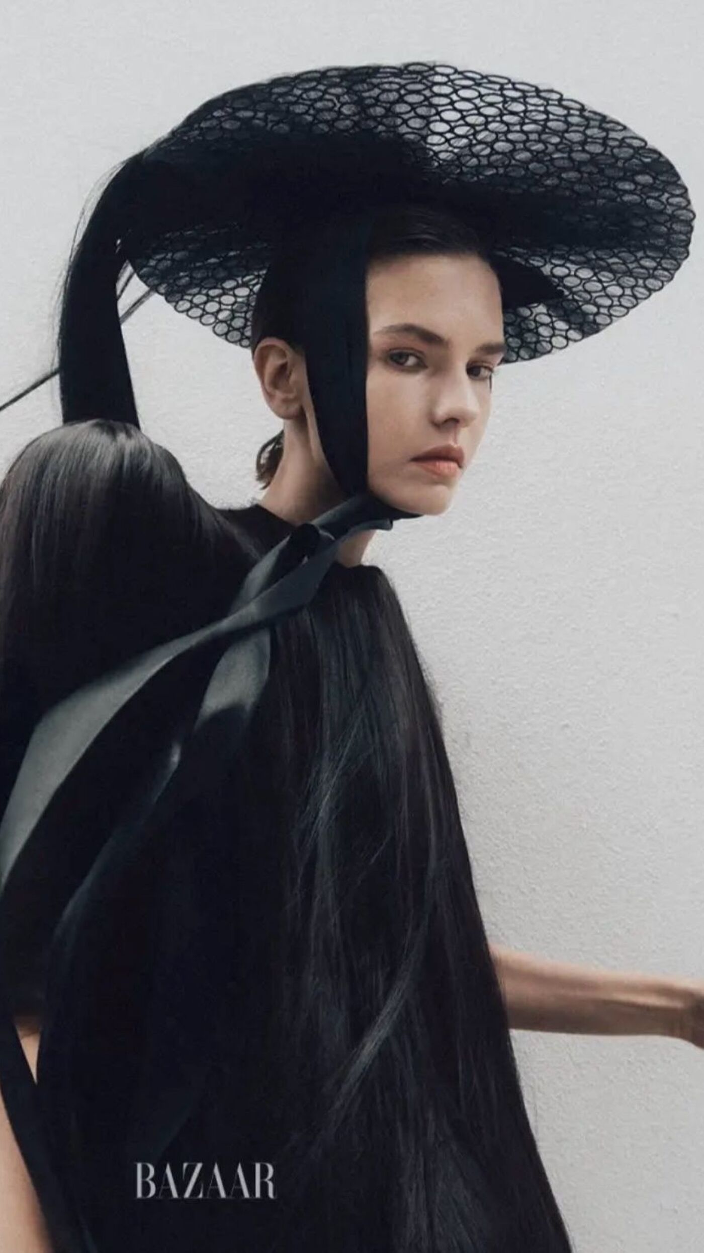 A woman wearing an extravagant black hat and a long, flowing dress against a white background. The image features a BAZAAR logo in the bottom right corner.