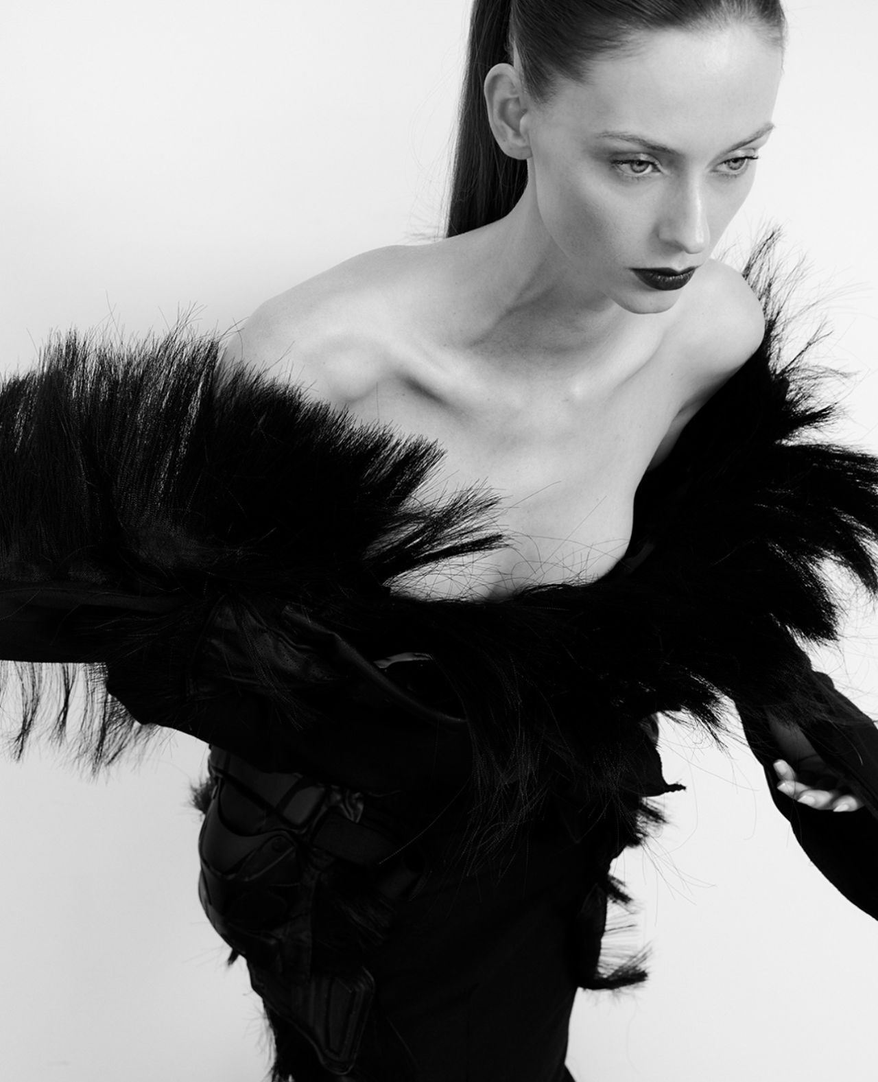 A woman in a black feathered dress, with her hair in a high ponytail, looks to the right with dramatic makeup.