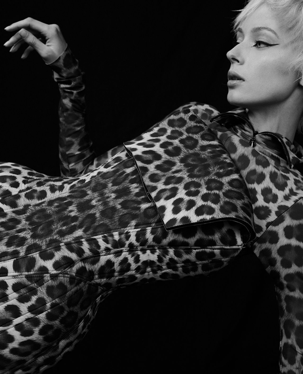 A woman with short hair and a leopard print dress poses against a black background. Her makeup features a dramatic winged eyeliner.