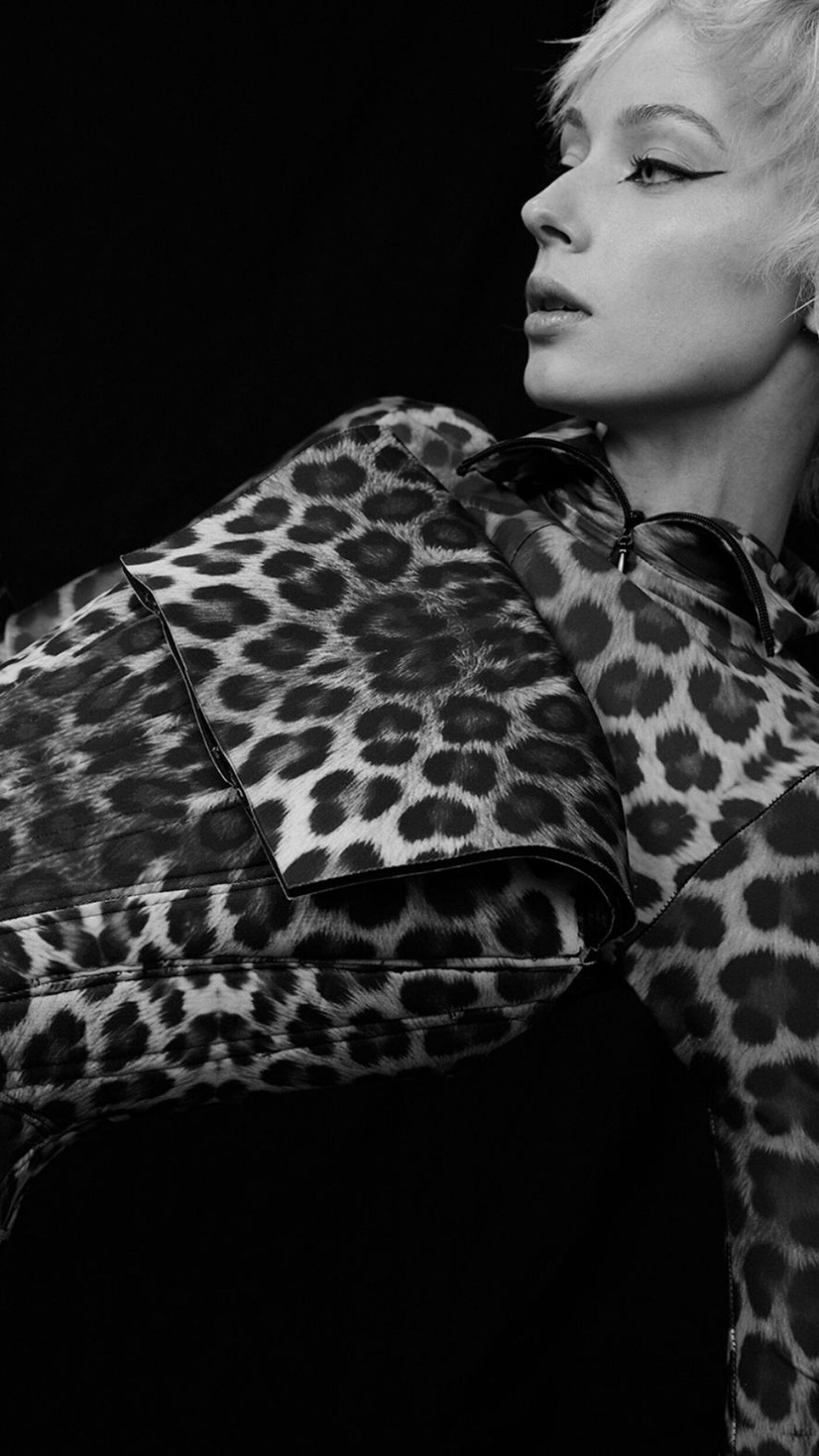 A woman with short hair and a leopard print dress poses against a black background. Her makeup features a dramatic winged eyeliner.