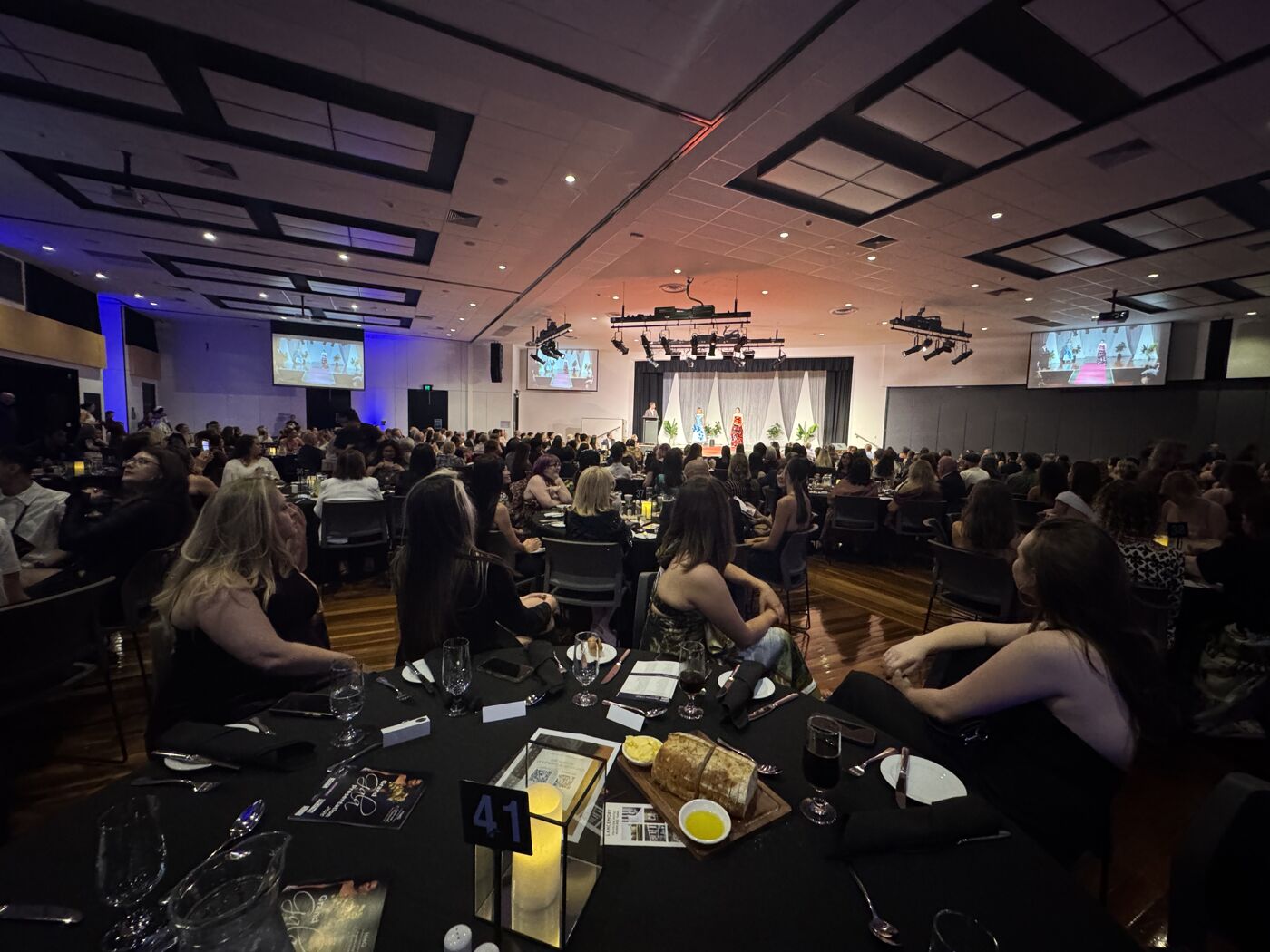 The image shows a large indoor event with many people seated at round tables, facing a stage with a presentation. The atmosphere appears to be formal, possibly a conference or awards ceremony.