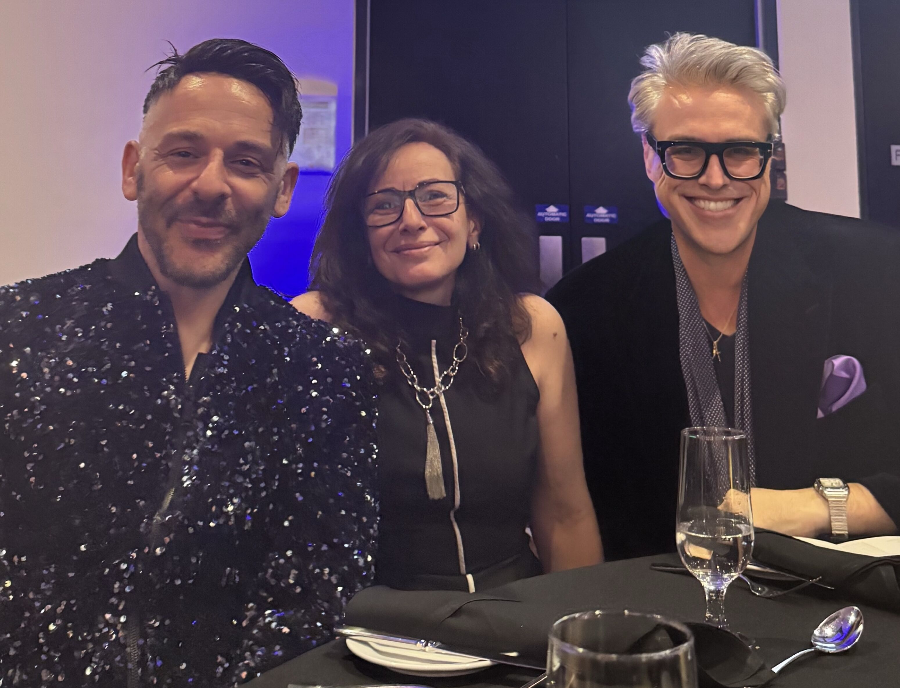 Three people are posing for a picture at a table. The person on the left is wearing a sparkly black shirt, the person in the middle is wearing glasses and a black dress, and the person on the right is wearing a black jacket and glasses.