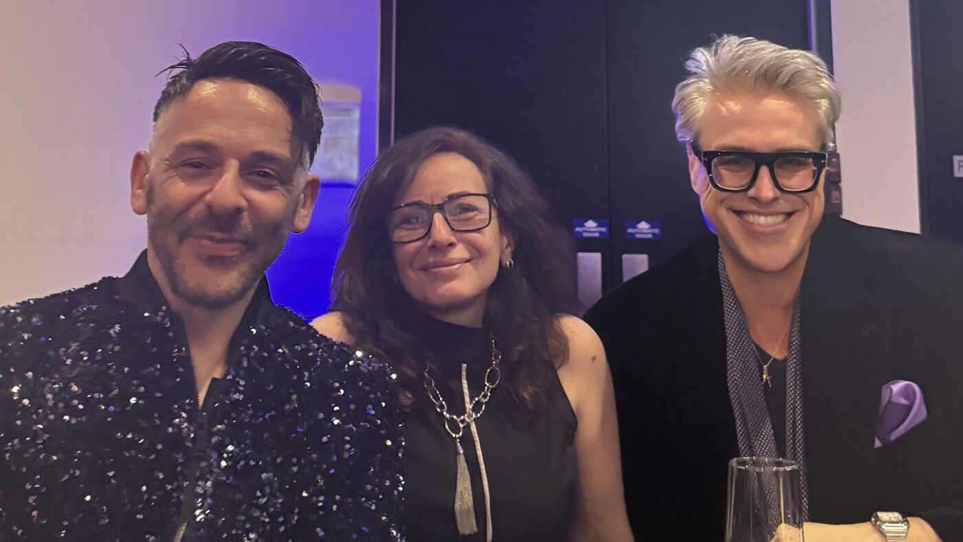 Three people are posing for a picture at a table. The person on the left is wearing a sparkly black shirt, the person in the middle is wearing glasses and a black dress, and the person on the right is wearing a black jacket and glasses.