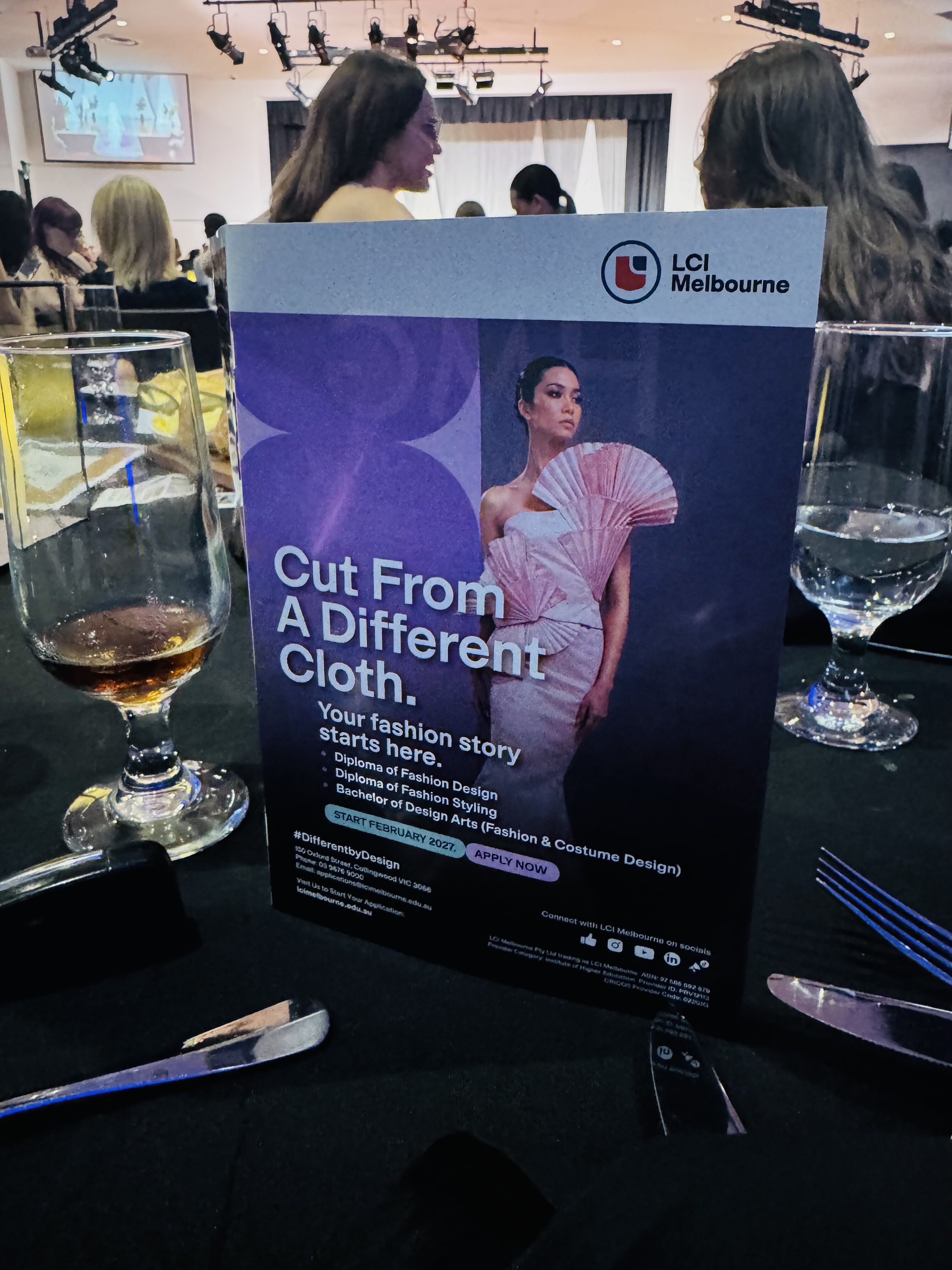 This image shows a table tent card from LCI Melbourne, a fashion design school, with the tagline "Cut From A Different Cloth. Your fashion story starts here." It features a model in a stylish outfit.