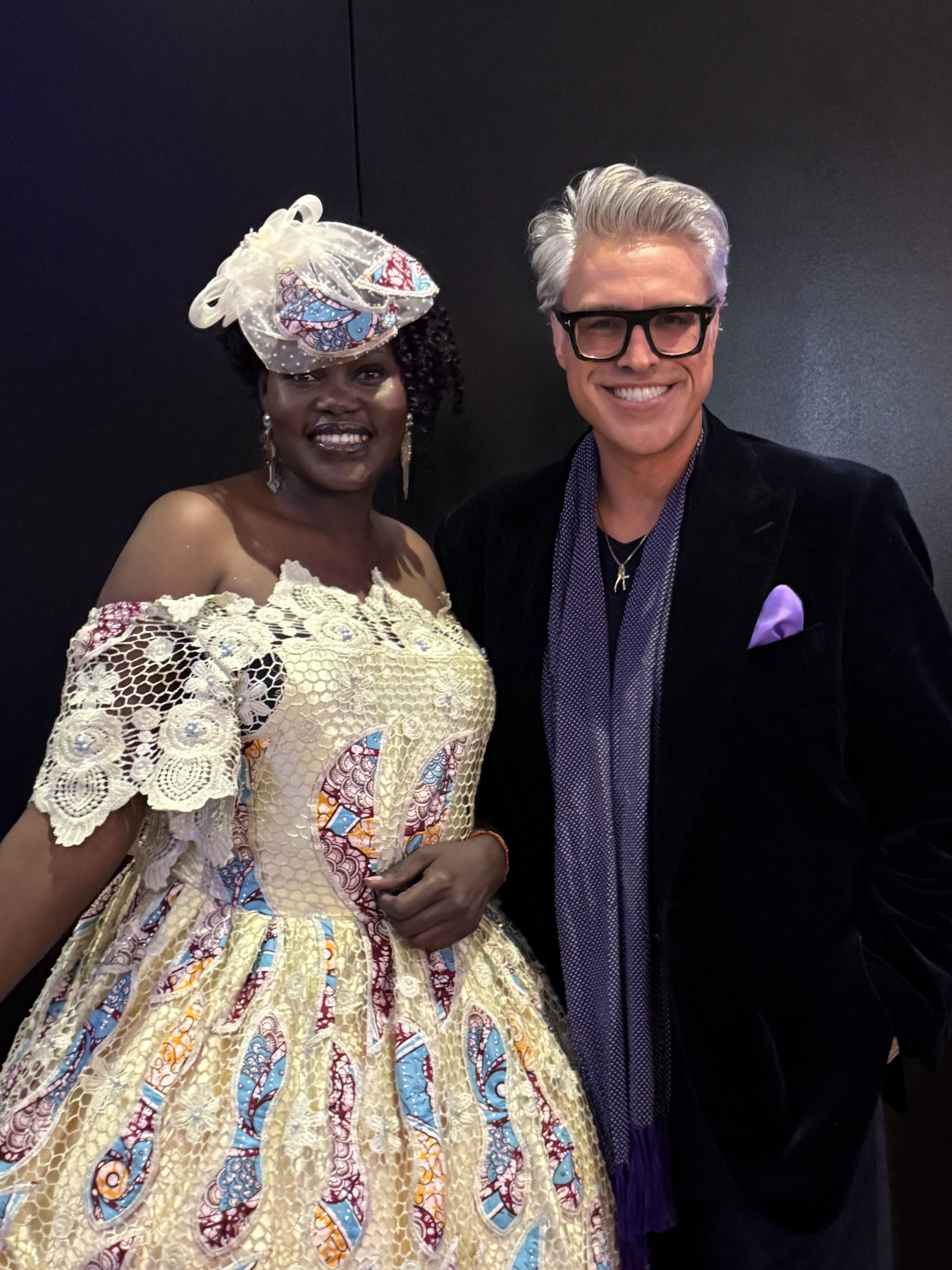 This image features a woman in a vibrant, off-the-shoulder dress and an ornate hat, posing next to a man in a dark suit with a purple scarf and pocket square.