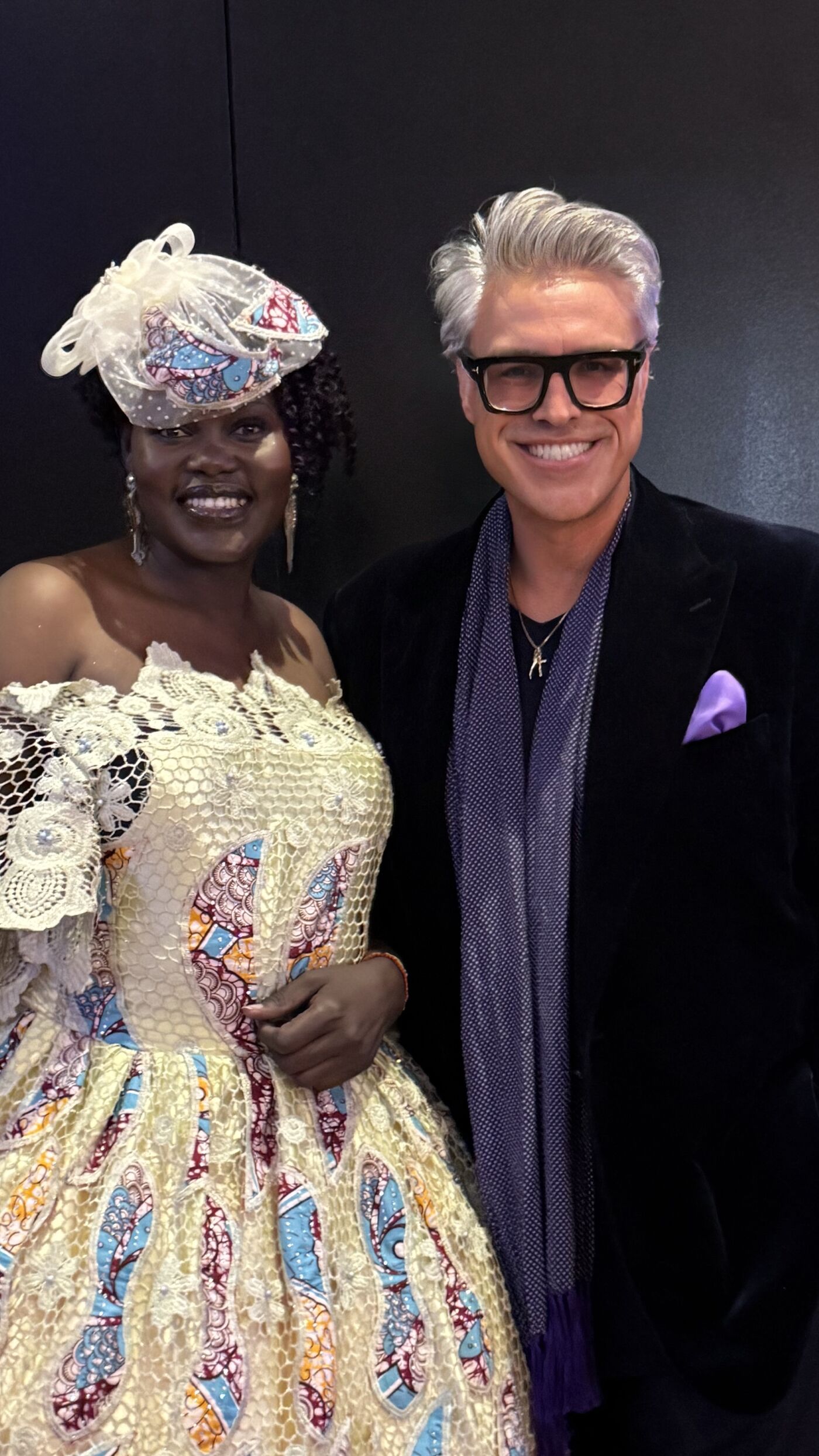 This image features a woman in a vibrant, off-the-shoulder dress and an ornate hat, posing next to a man in a dark suit with a purple scarf and pocket square.