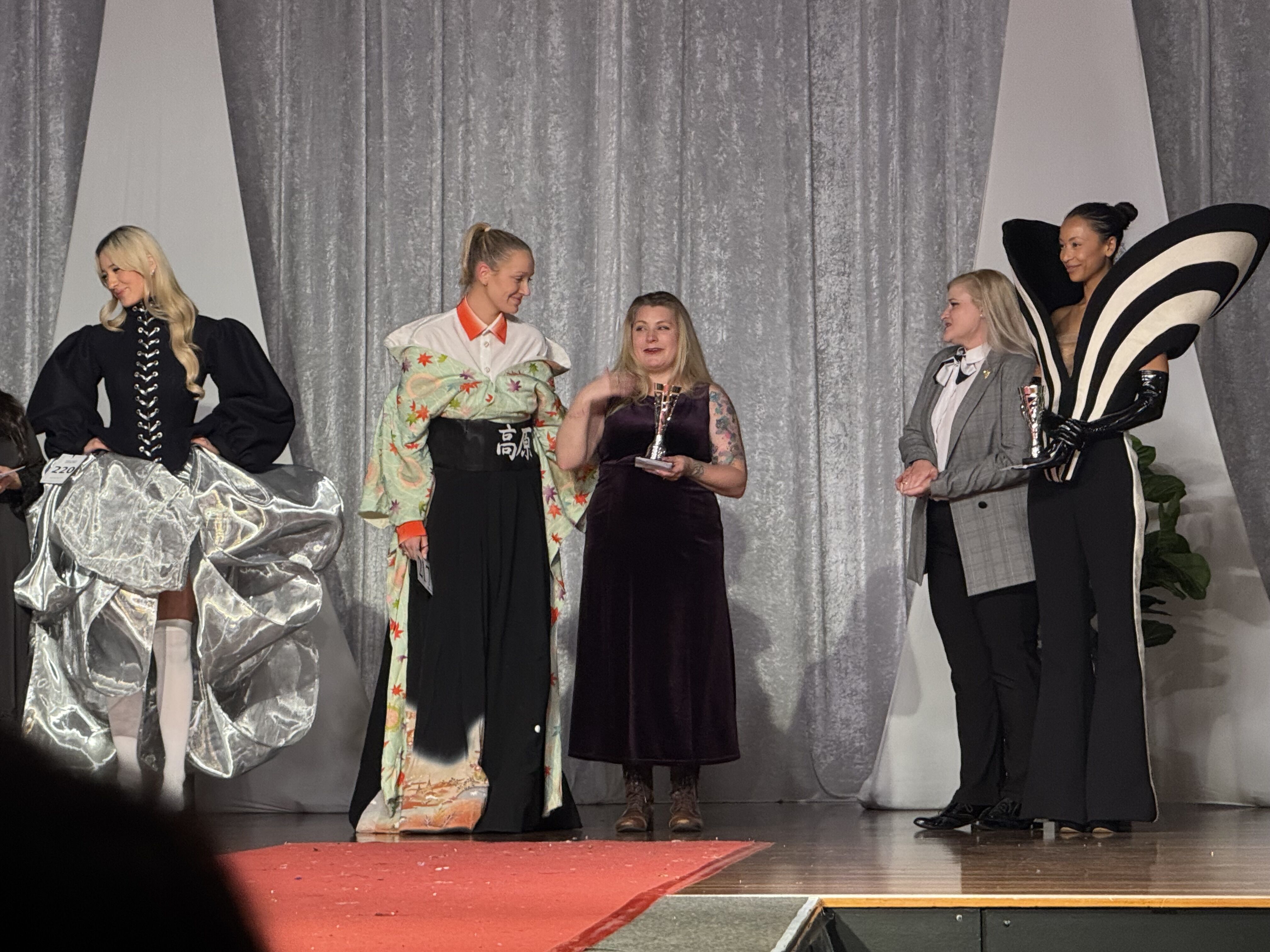 A group of people are on a stage, some are wearing elaborate costumes, and one person is holding a trophy, possibly at an awards ceremony or fashion show.