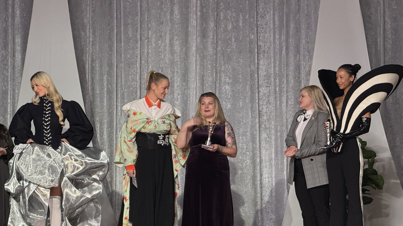 A group of people are on a stage, some are wearing elaborate costumes, and one person is holding a trophy, possibly at an awards ceremony or fashion show.