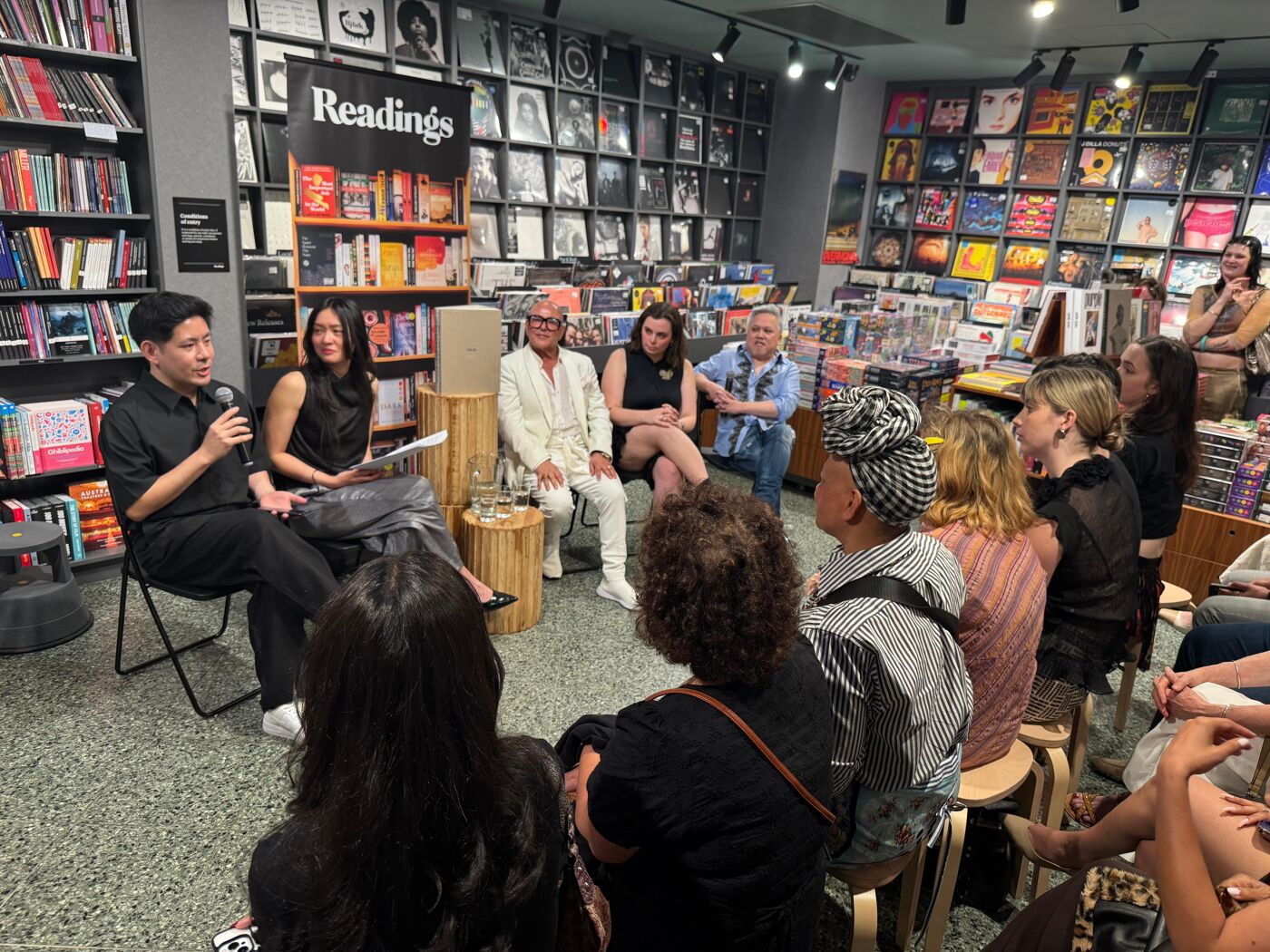 A group of people are gathered in a bookstore, listening to a speaker at a panel discussion. The room is filled with books and records, creating a cozy and intellectual atmosphere.