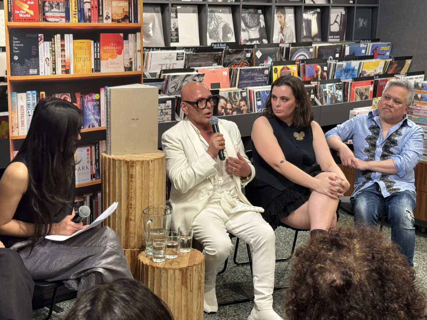 A panel discussion is taking place with four people. A man in a white suit is speaking into a microphone, with two women and another man seated and listening.