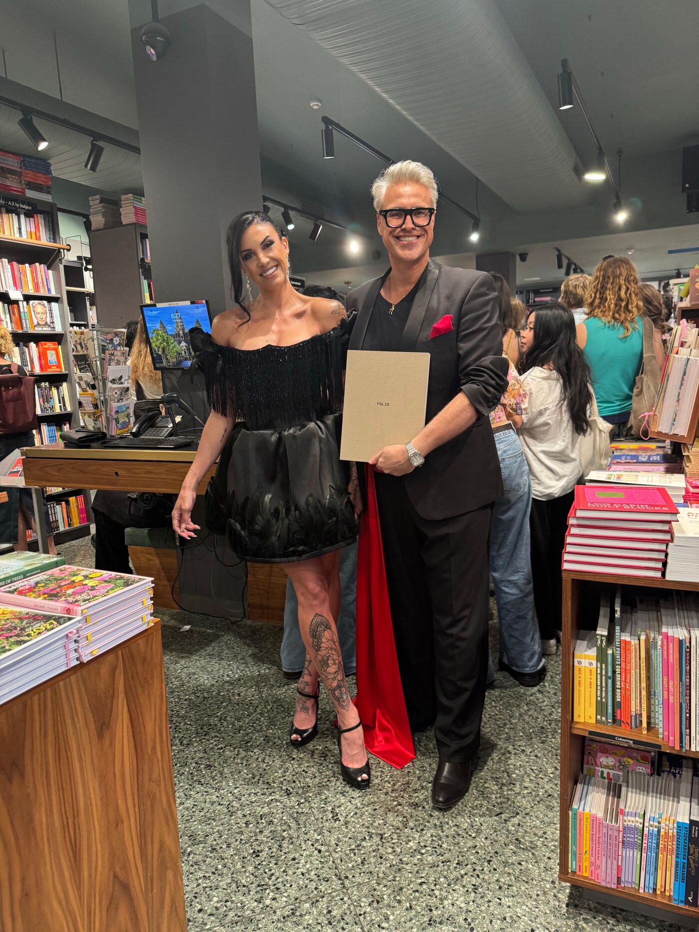 A woman in a black off-the-shoulder dress and a man in a black suit with a red cape are posing for a photo in a bookstore, holding a book.