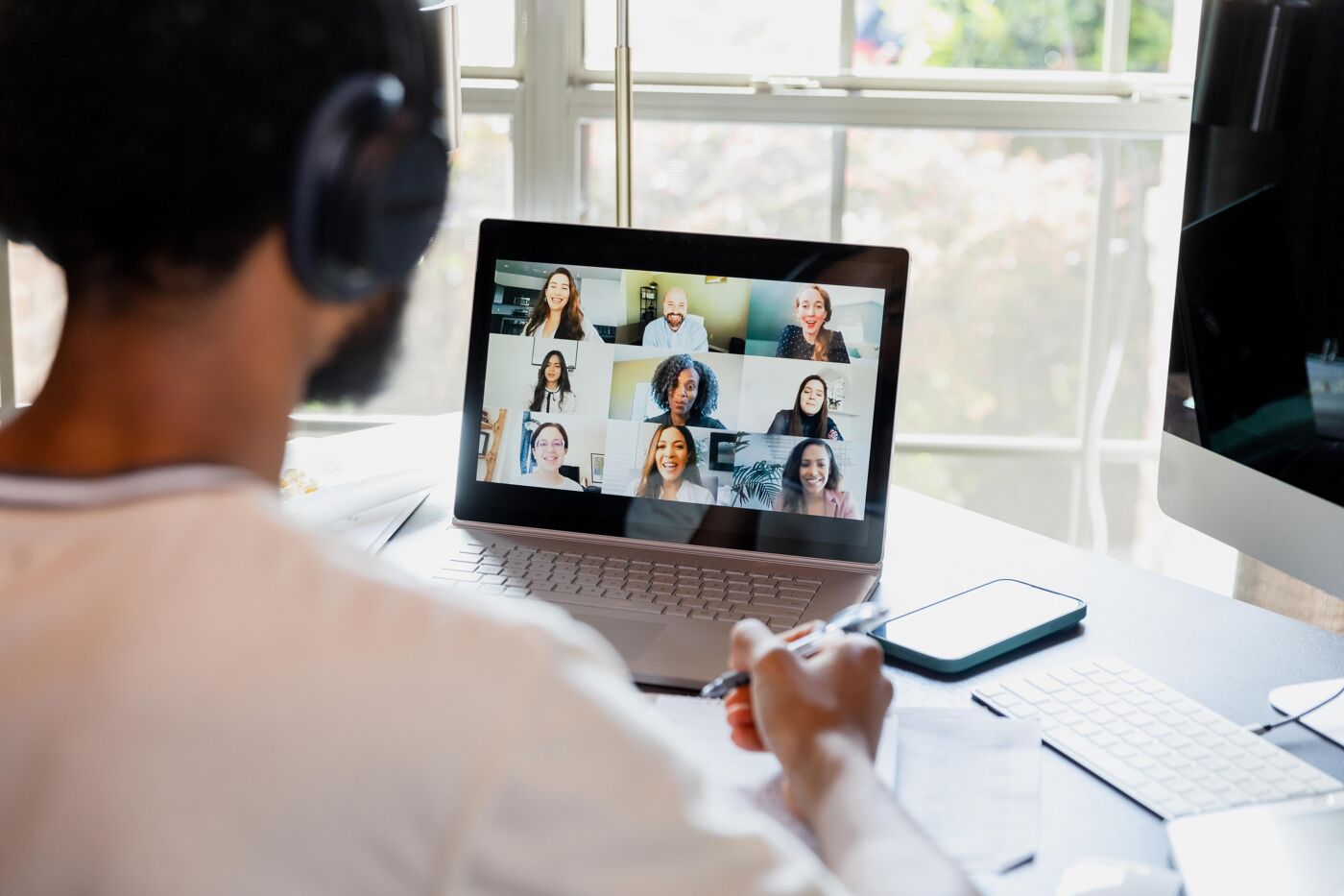 A person wearing headphones is participating in a video conference with multiple people on their laptop screen, taking notes.