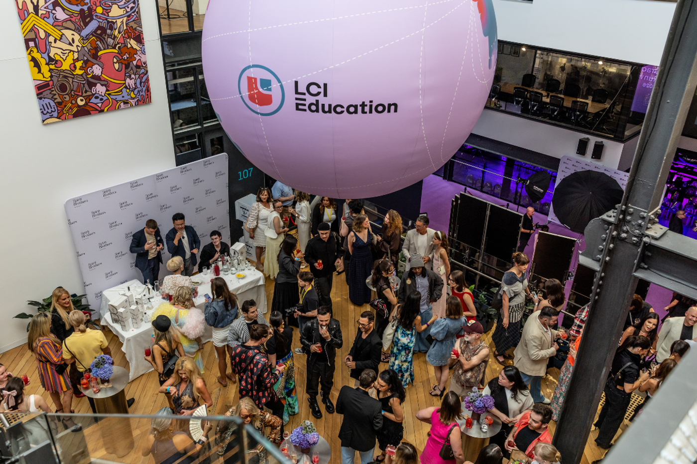 The image shows a crowded event with many people. A large balloon with the LCI Education logo hangs from the ceiling. People are socializing and enjoying themselves.