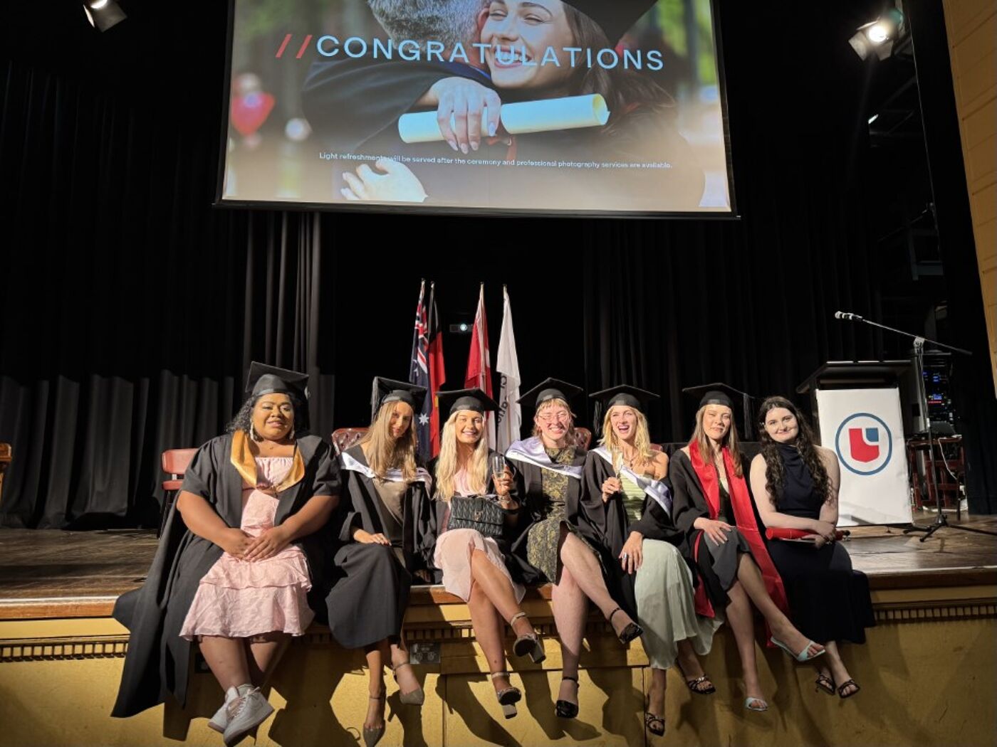 A group of female graduates are sitting on a stage in graduation gowns and caps, with a screen behind them displaying "Congratulations".