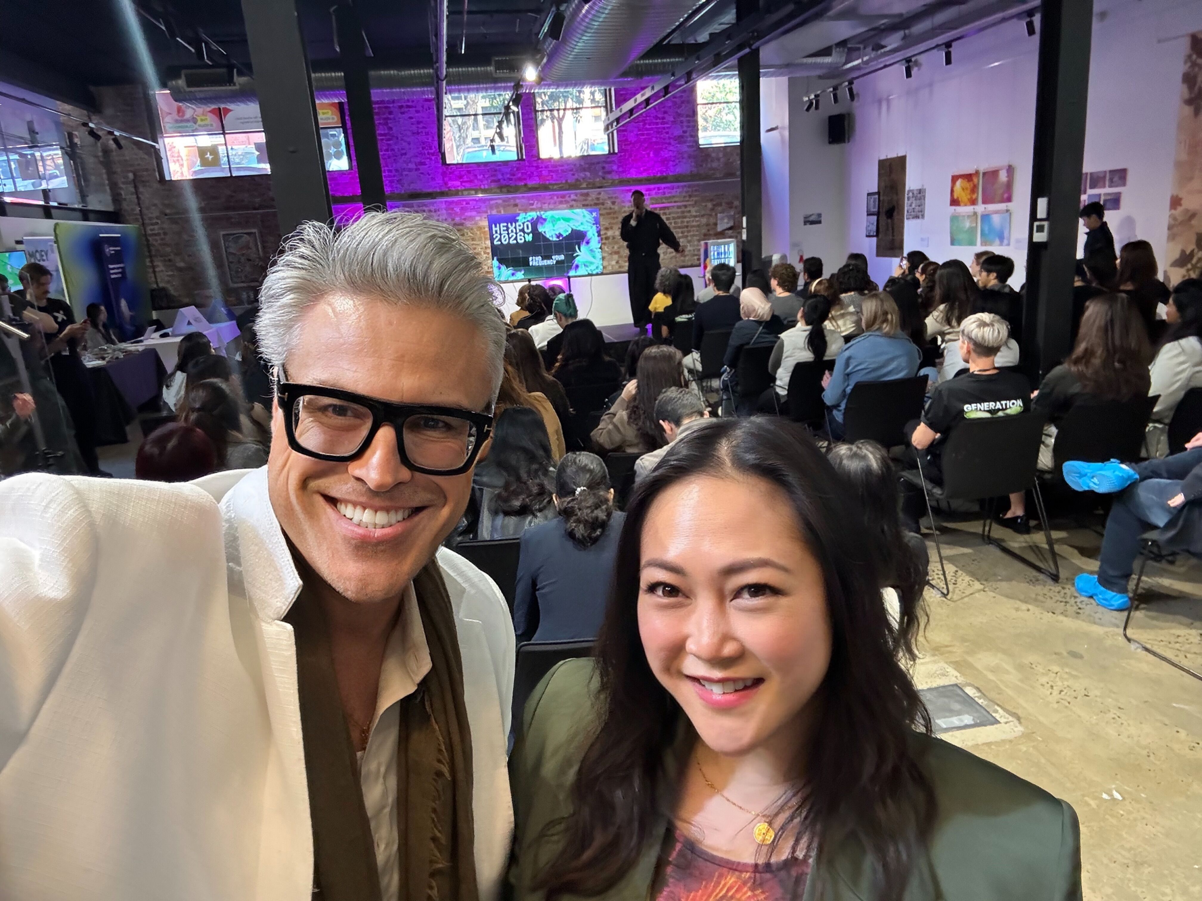 Two people smile at the camera in the foreground, with a crowd attending an event or conference in the background. The room has a modern feel with screens visible.