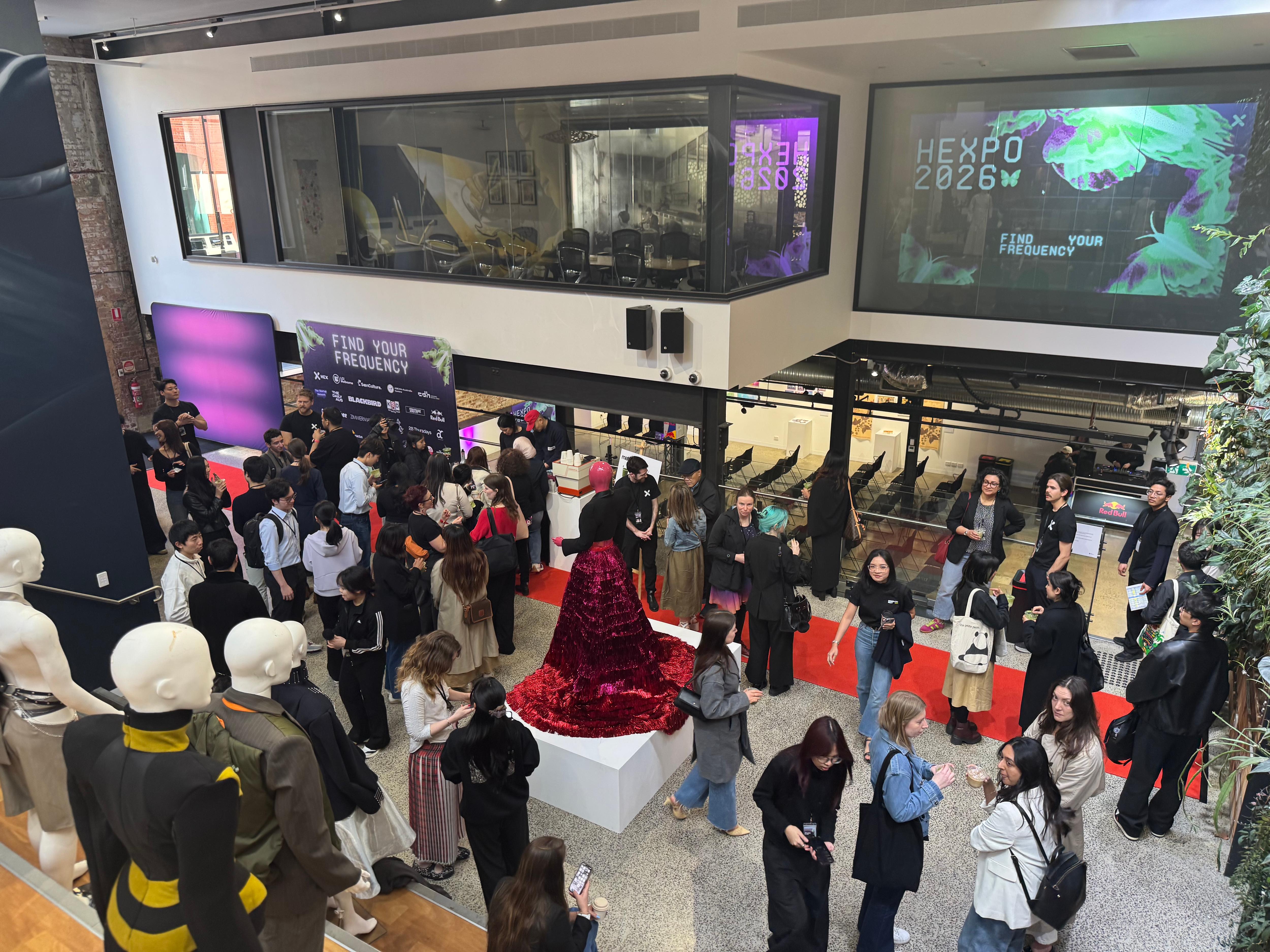 A crowd of people attends an indoor event, with screens displaying 'HEXP0 2026M' and 'FIND YOUR FREQUENCY'. A mannequin in a period costume stands in the foreground.