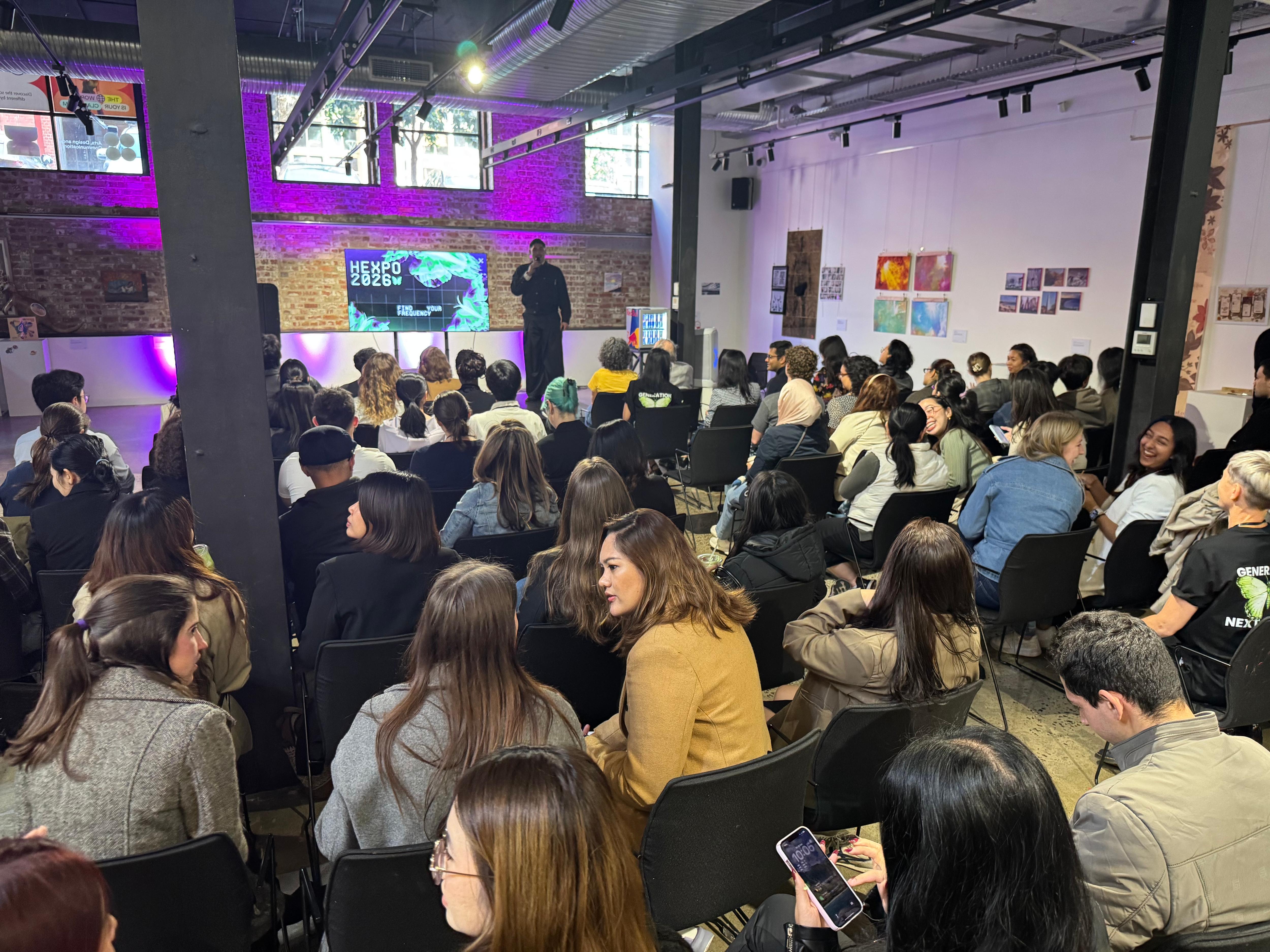 A crowd of people are attending a presentation or talk in an indoor space, with a speaker in front of a screen illuminated by purple lights.