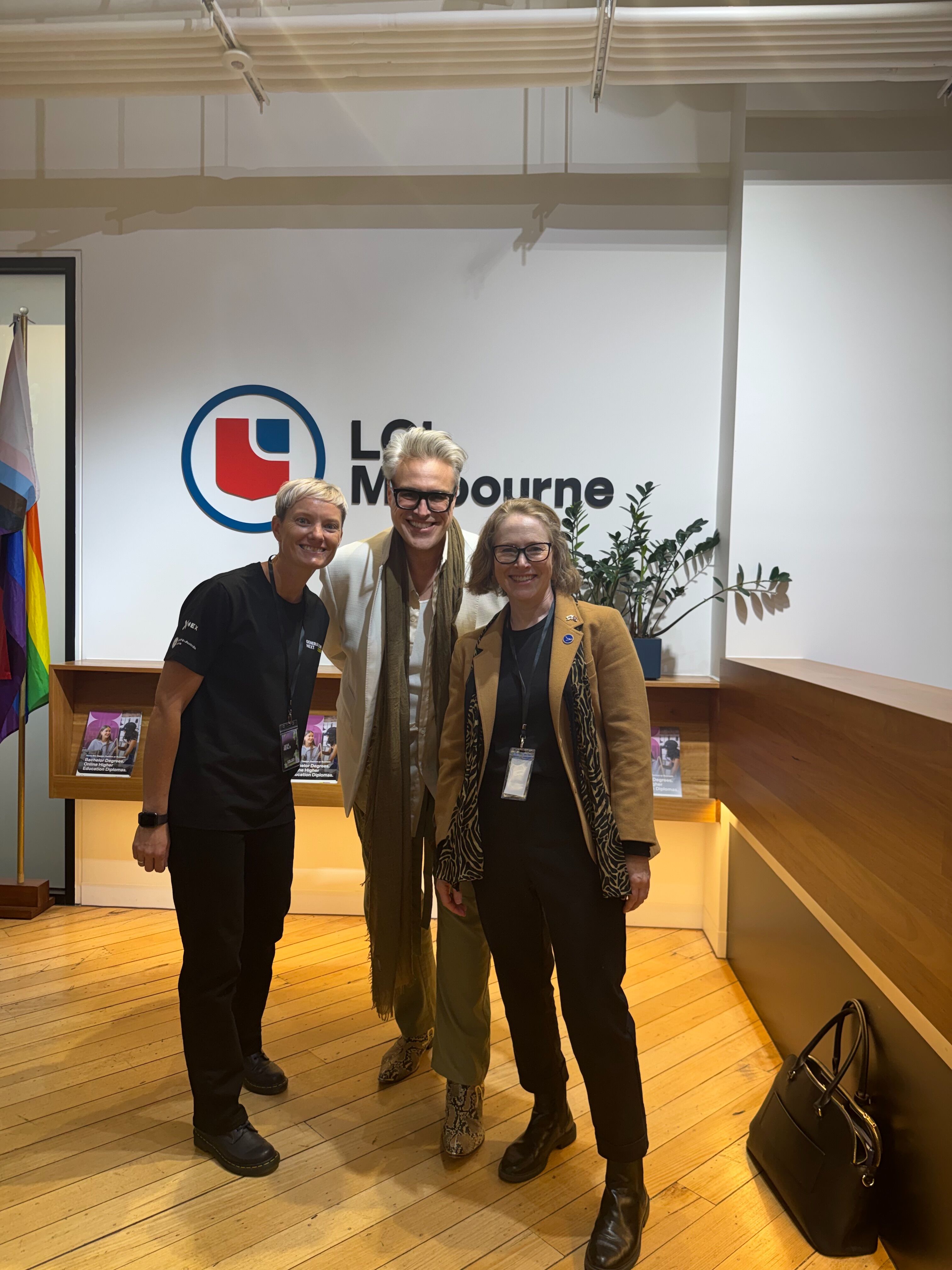 Three people smile at the camera in front of a logo that reads "LGBTI Melbourne". To the left, there is a rainbow flag.