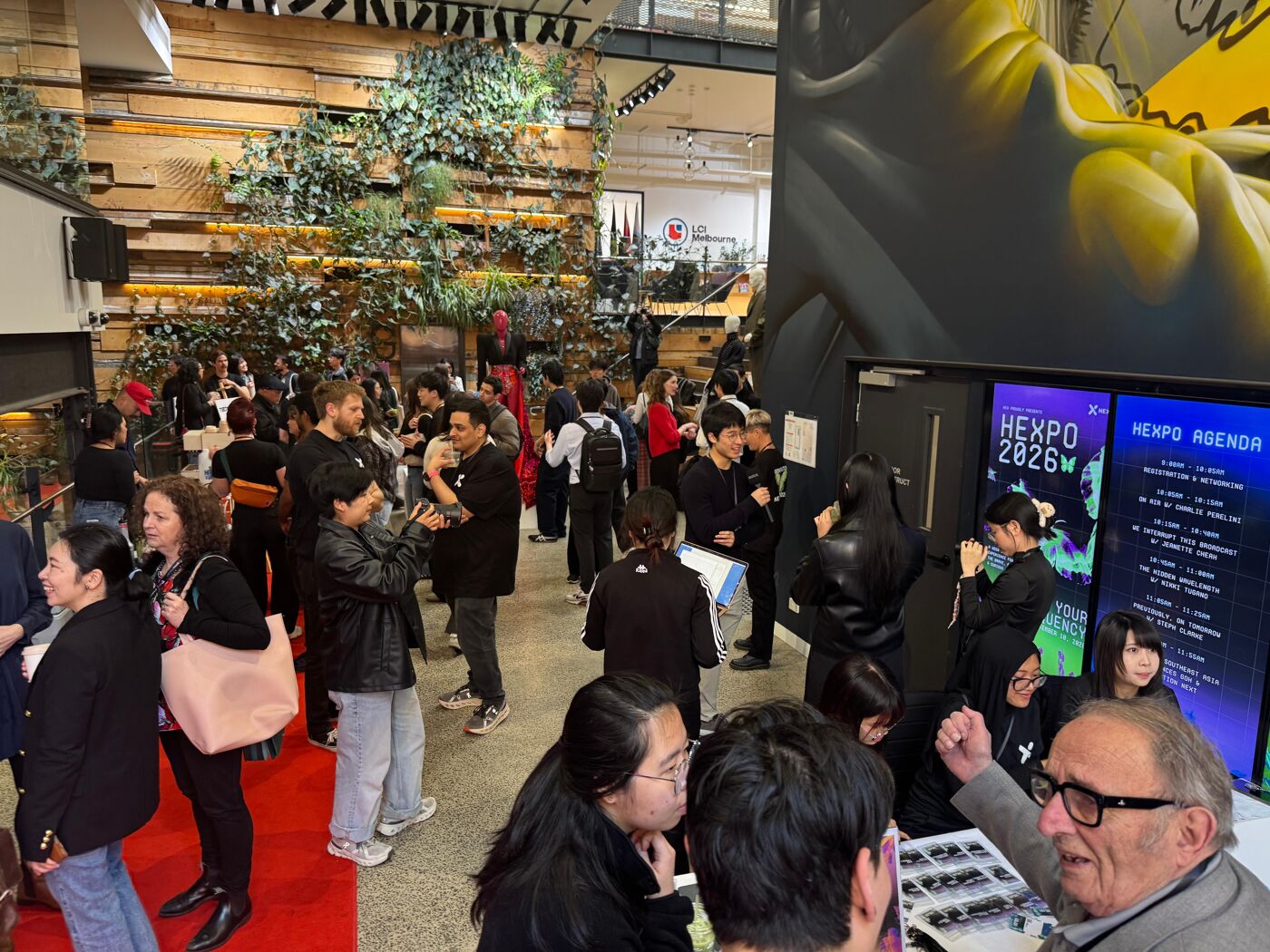 A crowd of people attends an indoor event, possibly a fair or exhibition, with green decorations and digital displays visible.