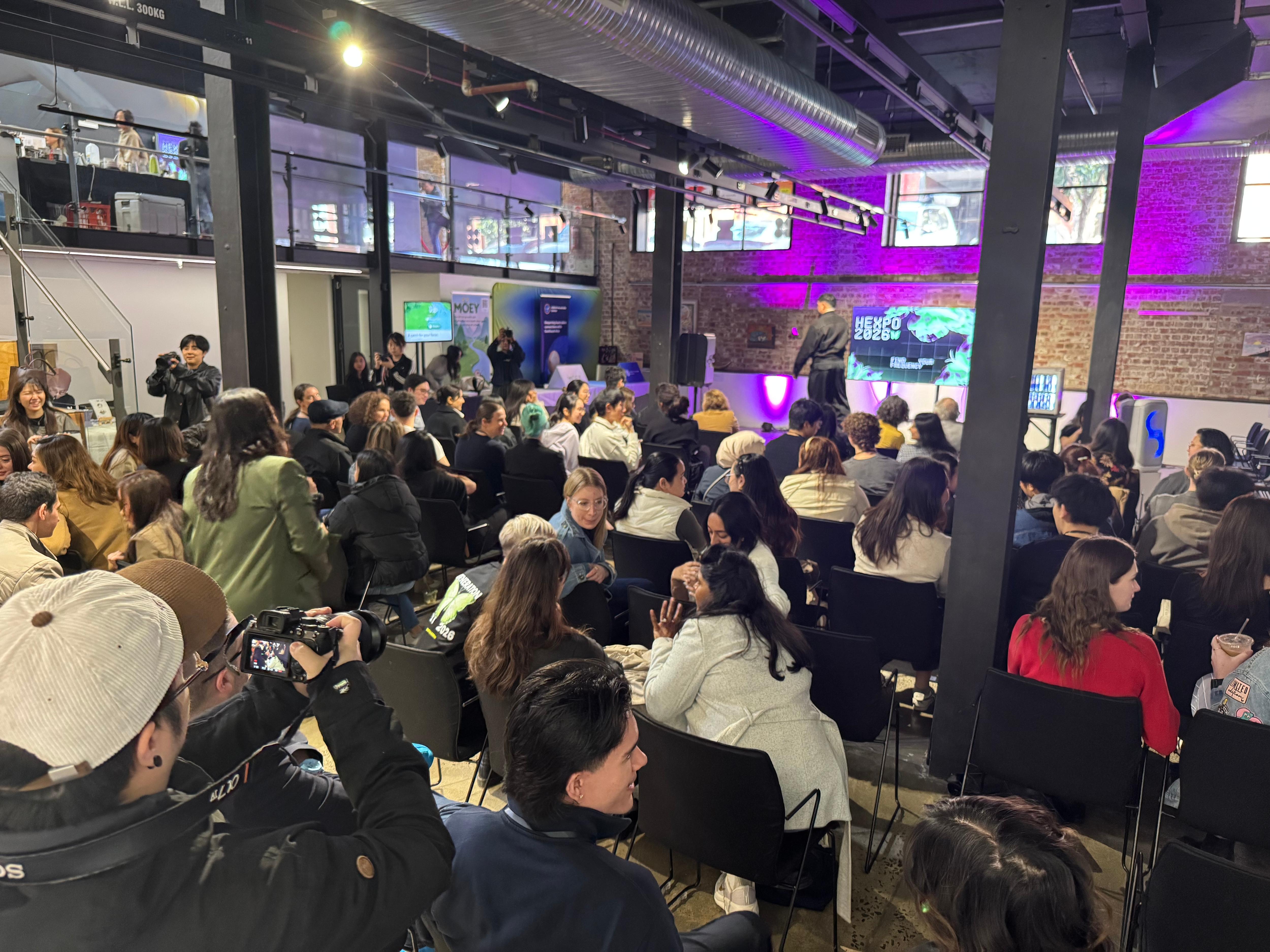 A crowd gathers in a room with purple lighting, watching a screen as a speaker presents. People are seated and standing, with some holding cameras.