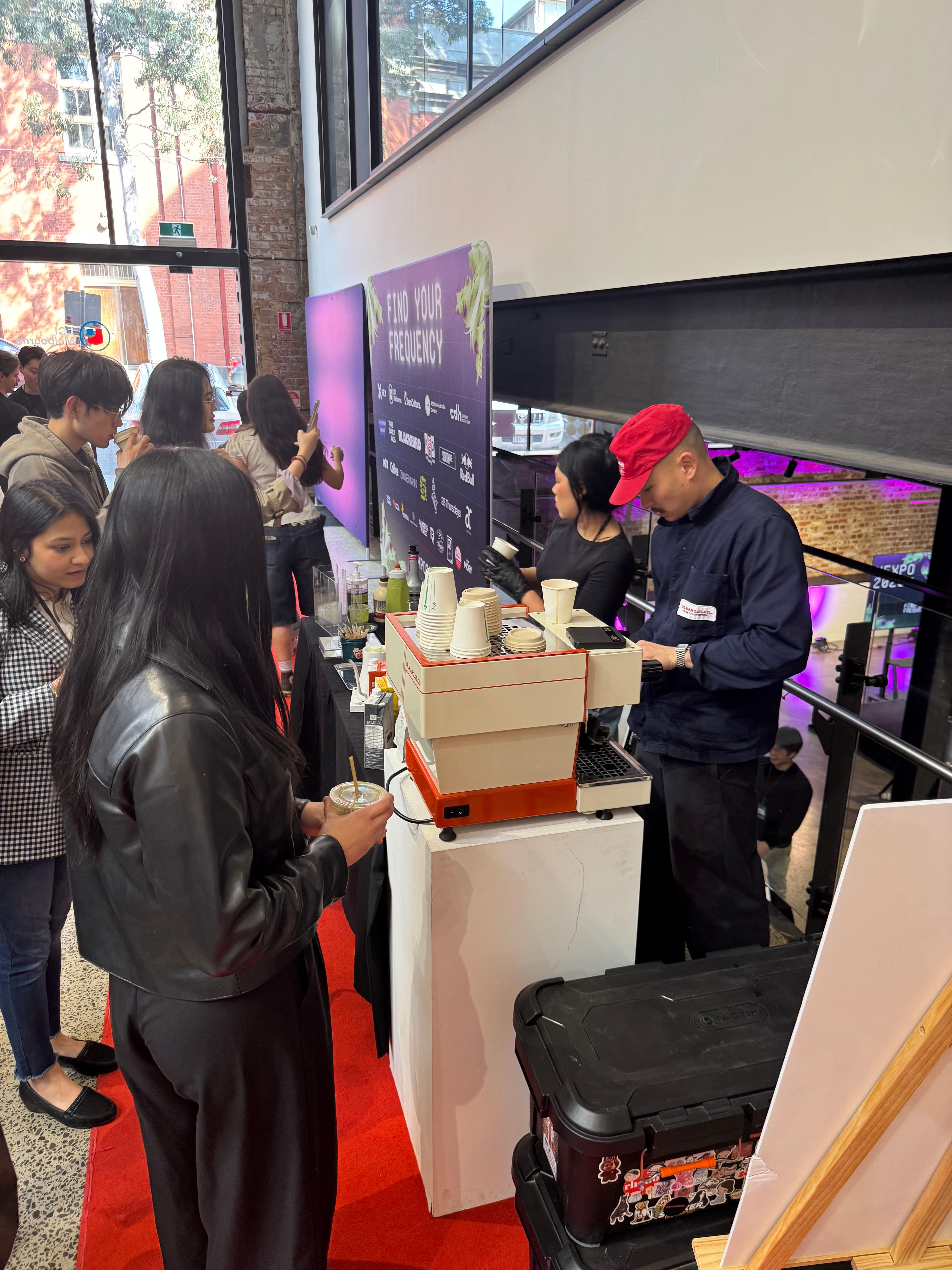 A man wearing a red cap is preparing coffee at a fair, with customers waiting around a counter with cups and equipment. Other people are observing in the background.