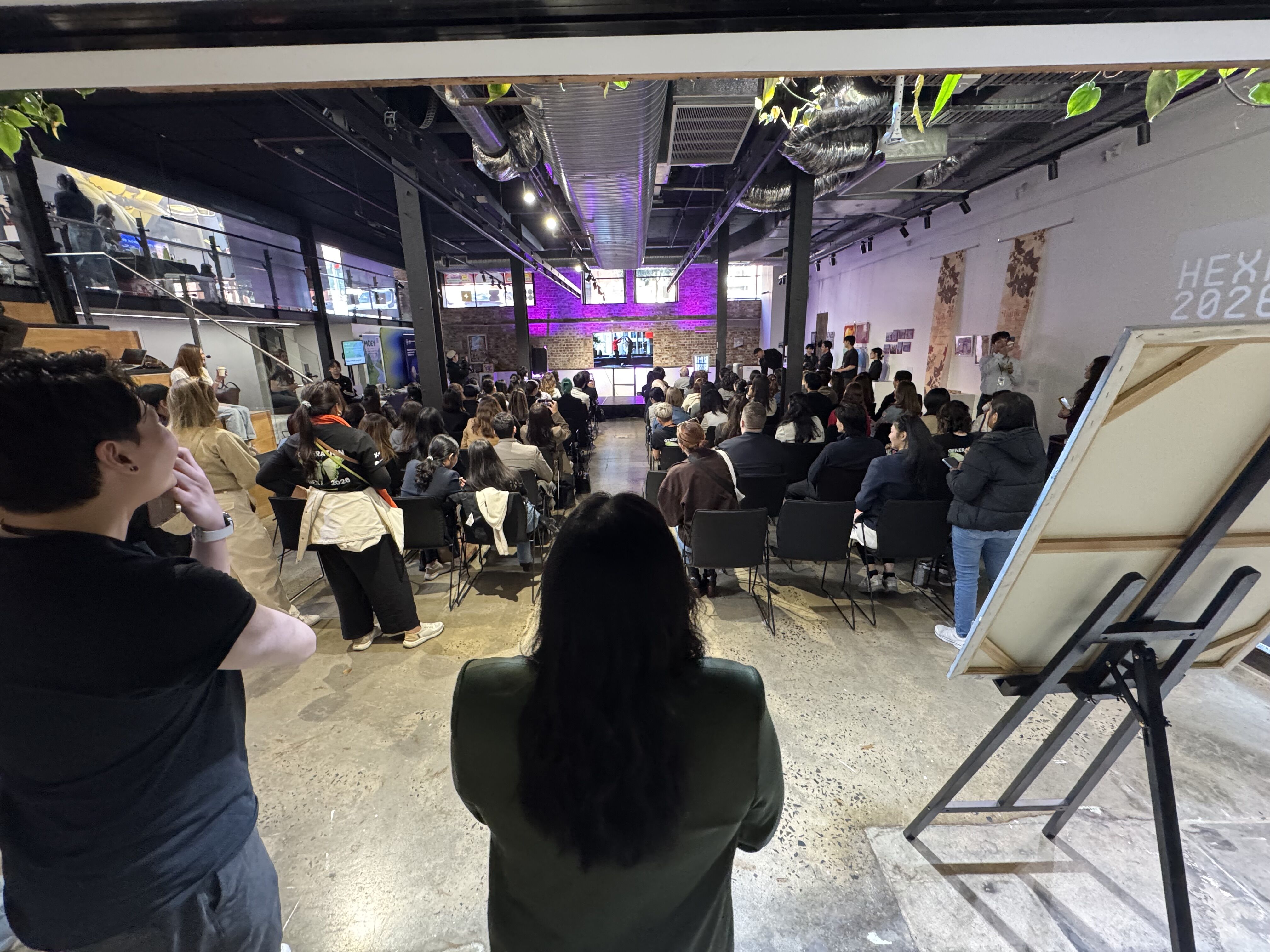 An indoor event is captured, showing a seated audience facing a stage with screens. A person stands in the foreground, observing the scene.