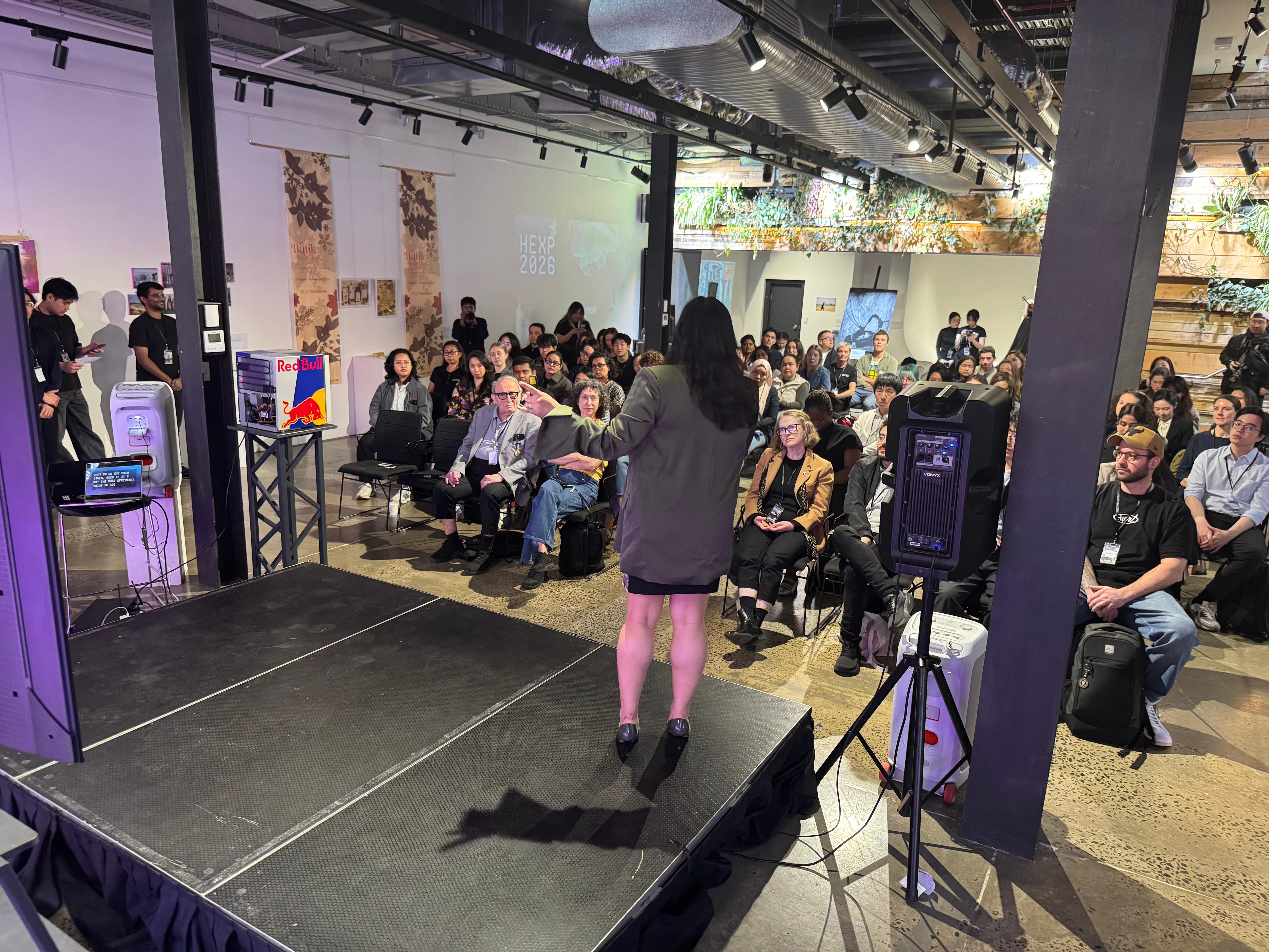 A woman speaks to a seated audience in an event space featuring stage lighting and plant decor.