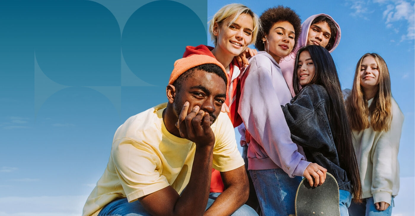 A diverse group of smiling young people, ranging from teens to young adults, pose together under a clear blue sky, some with skateboards.