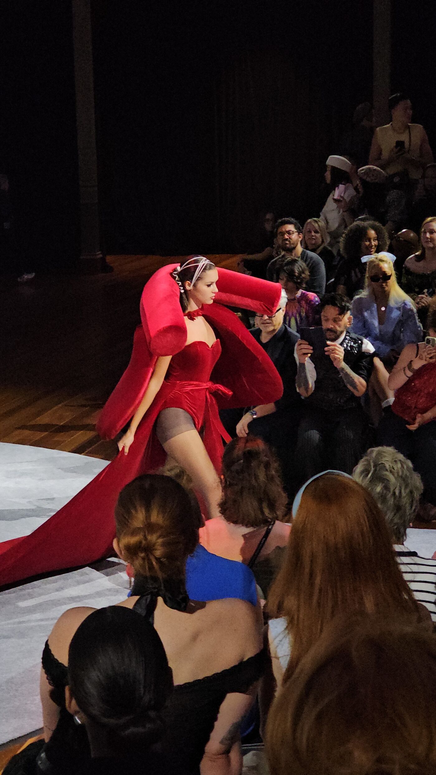 A model in a striking red dress walks the runway. The dress features a dramatic cape and a high slit, captivating the audience with its bold design.