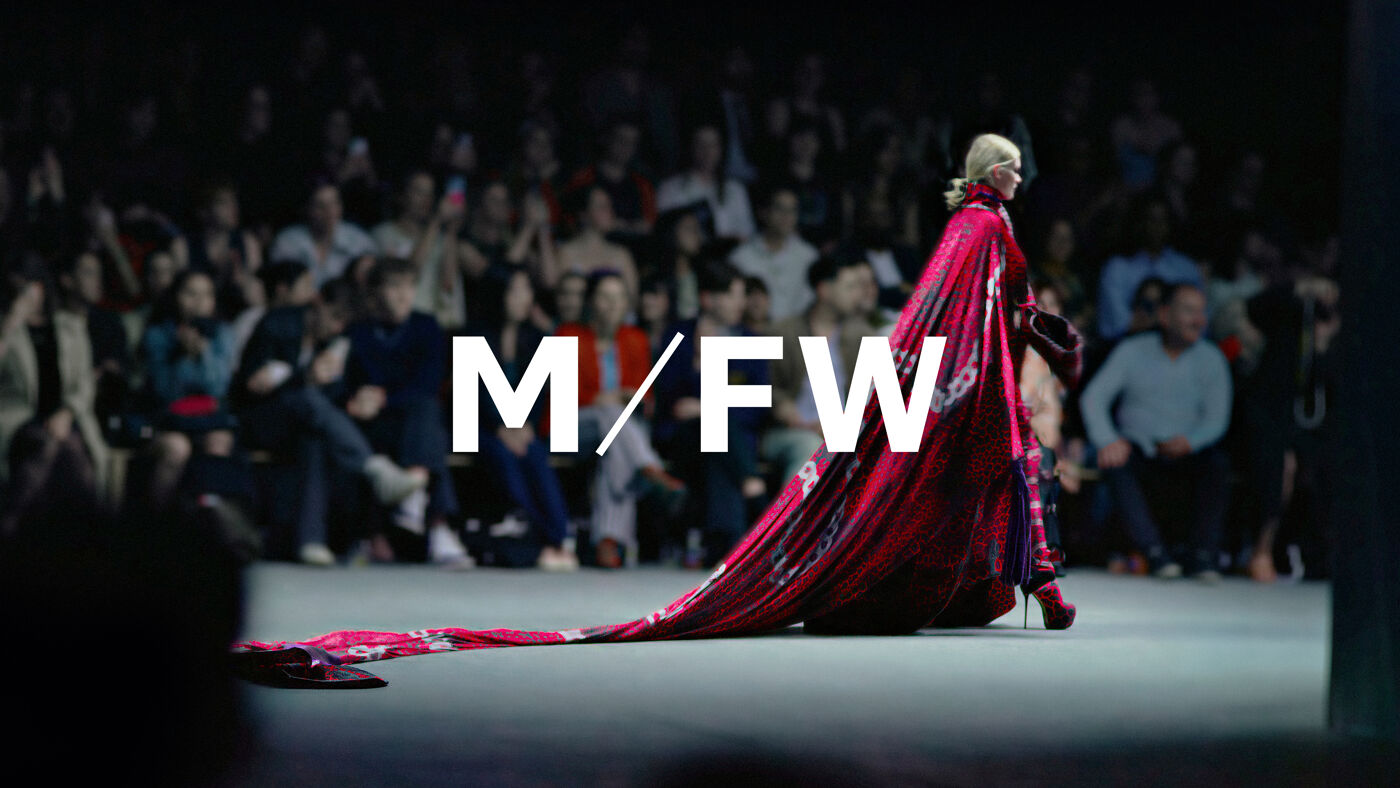 A woman walks down a runway in a dramatic, long red gown with an audience applauding in the background. The M/FW logo is visible.
