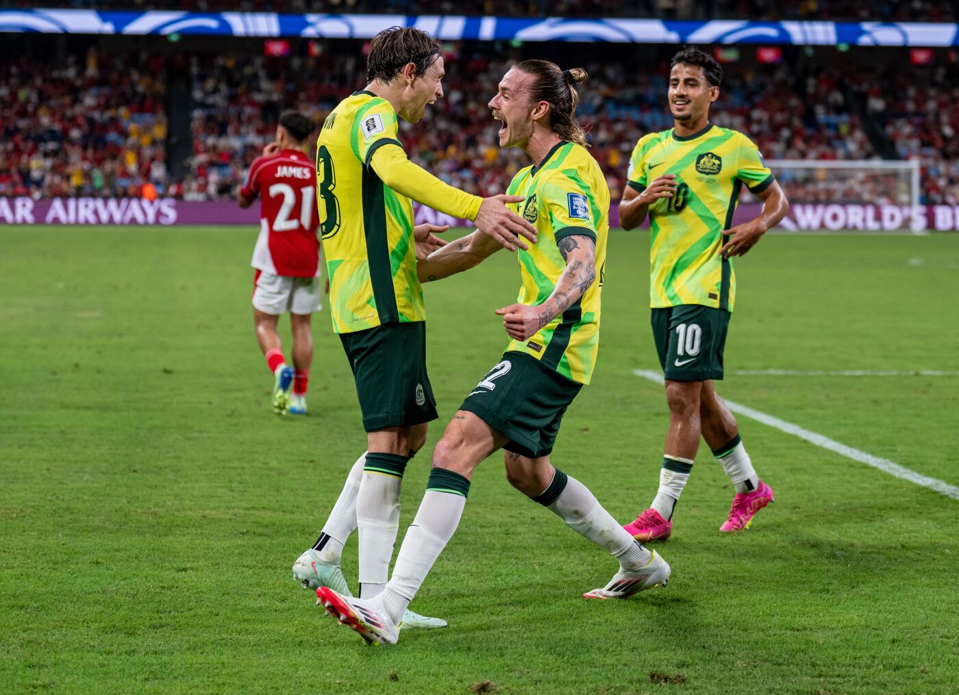 The image shows three soccer players in yellow and green uniforms on a green field, celebrating a goal. One player is jumping with his arms raised, while another is reaching out to hug him. A third player stands nearby with a smile.