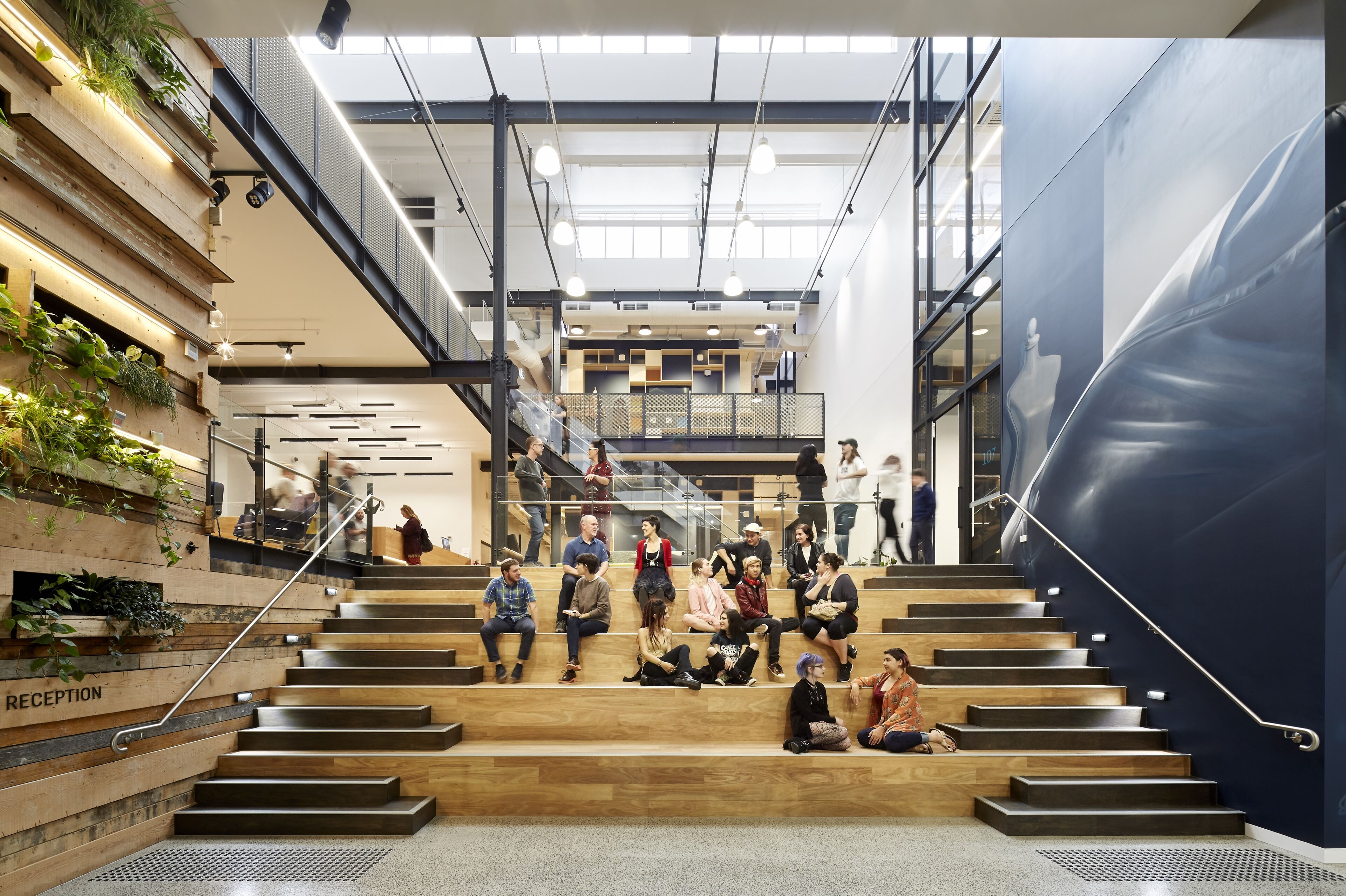 Students and staff engage in casual conversation on the amphitheater stairs of a modern atrium at LCI Melbourne Campus, surrounded by greenery and urban design.