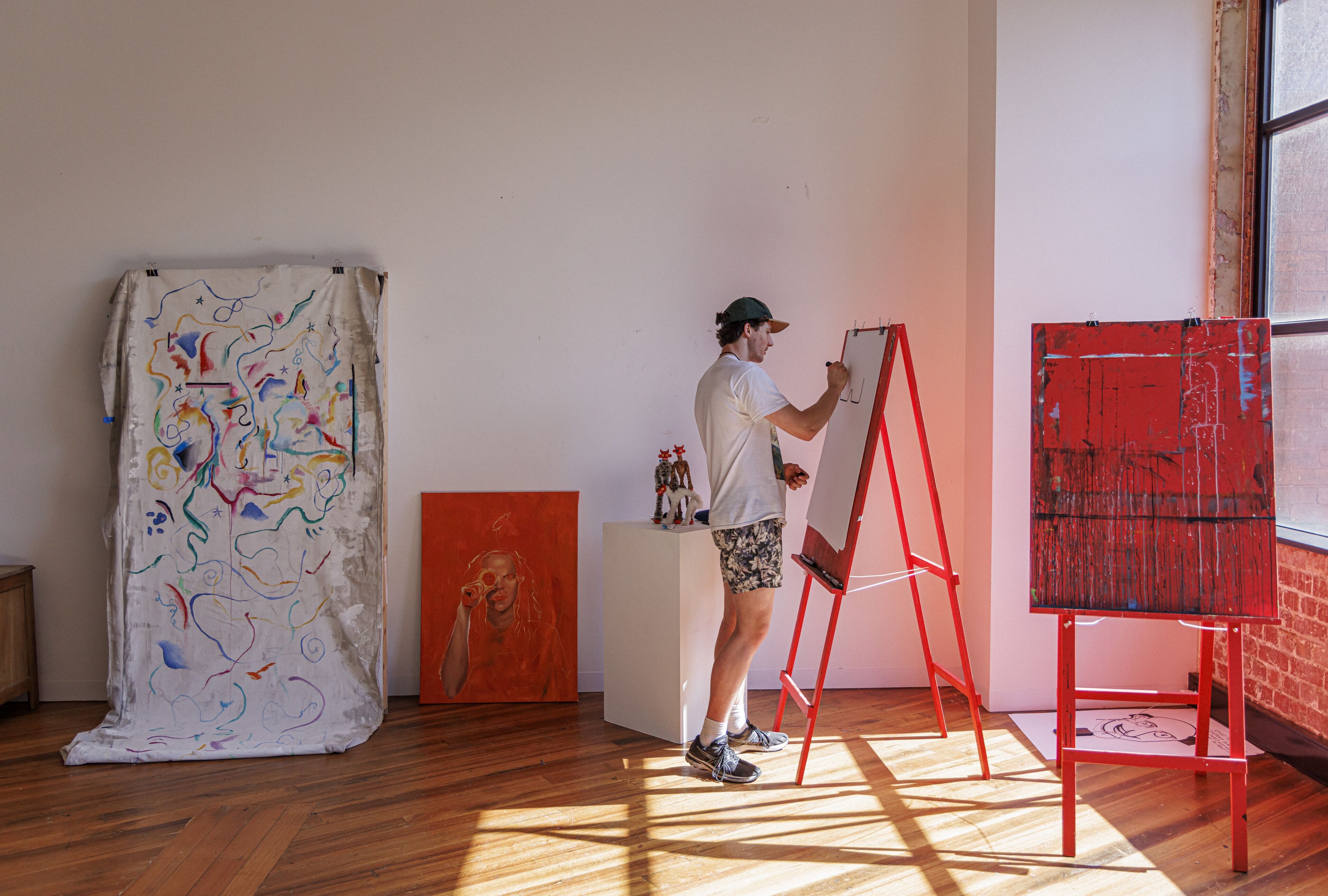 An artist sketches on an easel amid vibrant paintings in a sunlit studio.