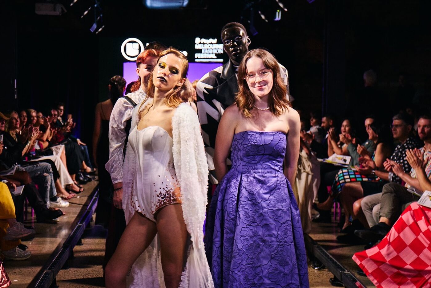 A runway show features models in various outfits, including a woman in a white sequined bodice and a purple dress, with spectators seated in the background.