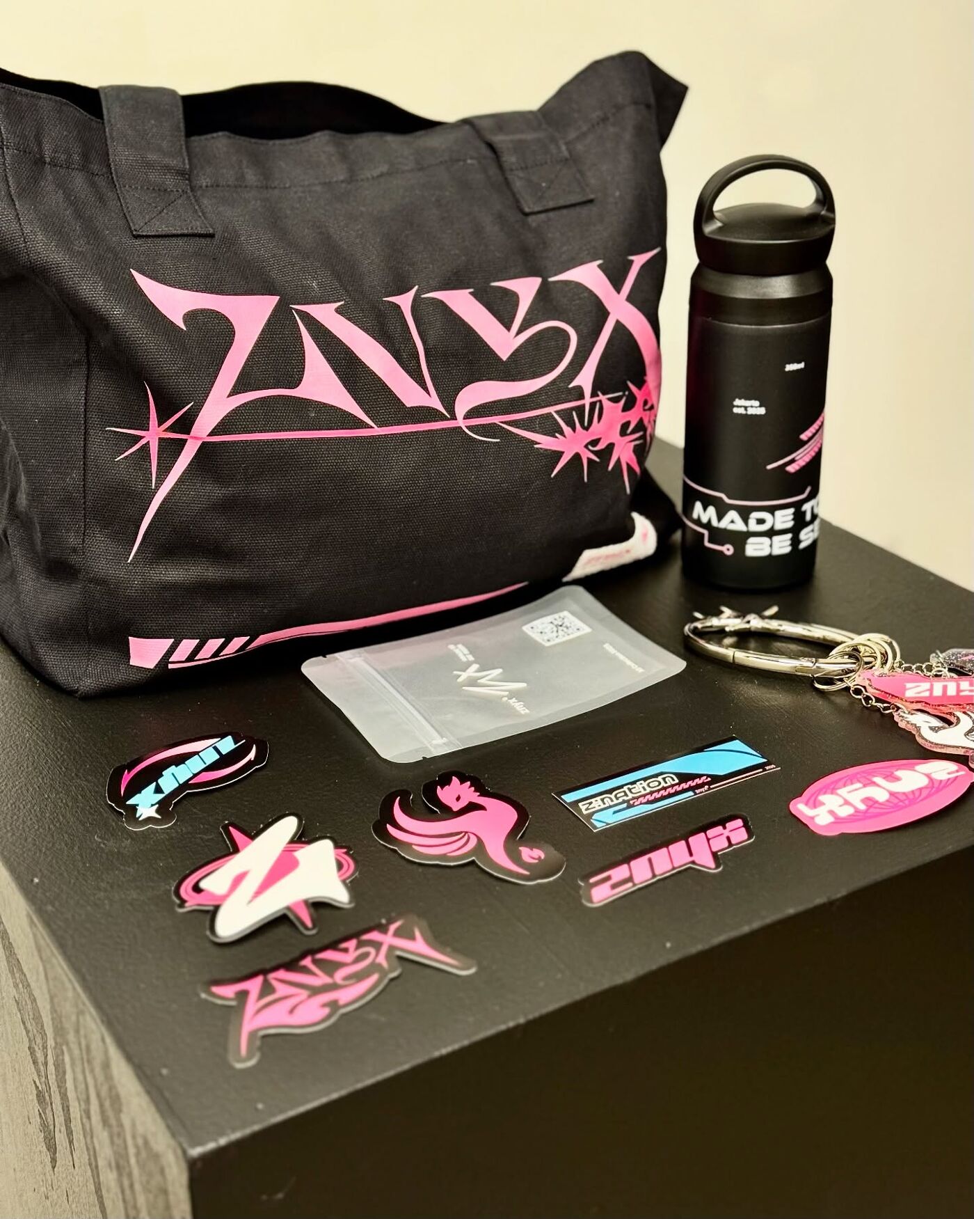 A collection of branded items including a black tote bag, water bottle, keychains, and colorful stickers with bold pink and blue designs, displayed on a black surface.
