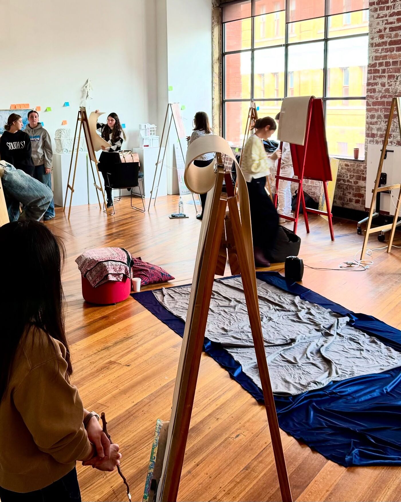 A bright art studio with students working on easels, sketching and painting. Fabrics are spread on the floor as part of the setup, creating a creative and collaborative atmosphere.