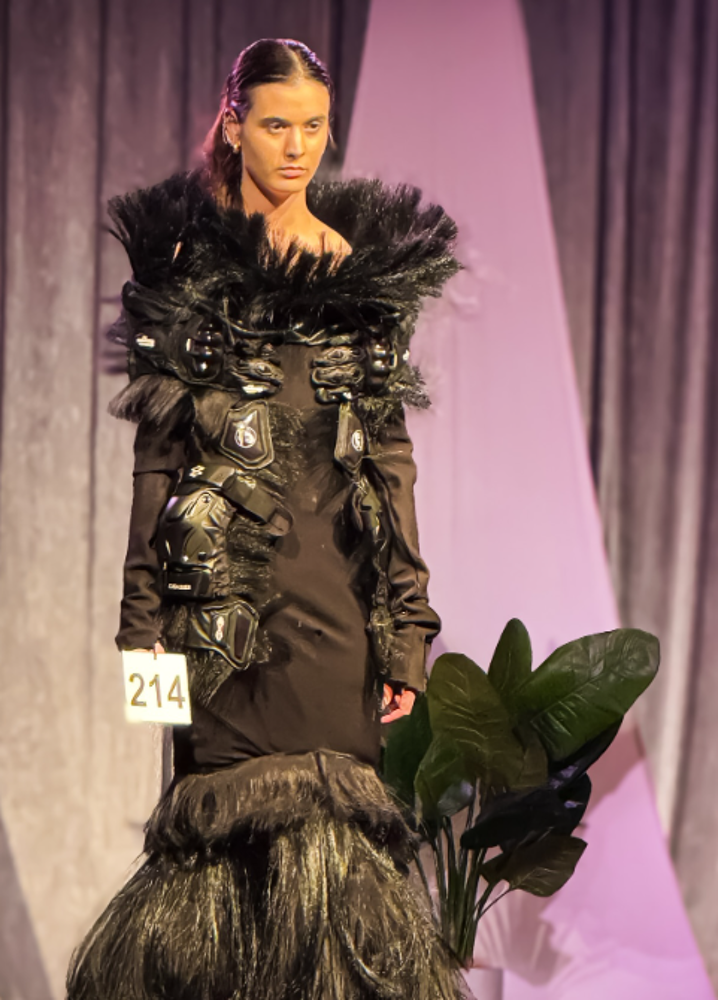 A model stands on a runway wearing a dark, avant-garde dress with elaborate feathered and textured embellishments. The dress features a fitted bodice and a voluminous skirt, creating a dramatic silhouette. The model's expression is serious, complementing the high-fashion aesthetic.
