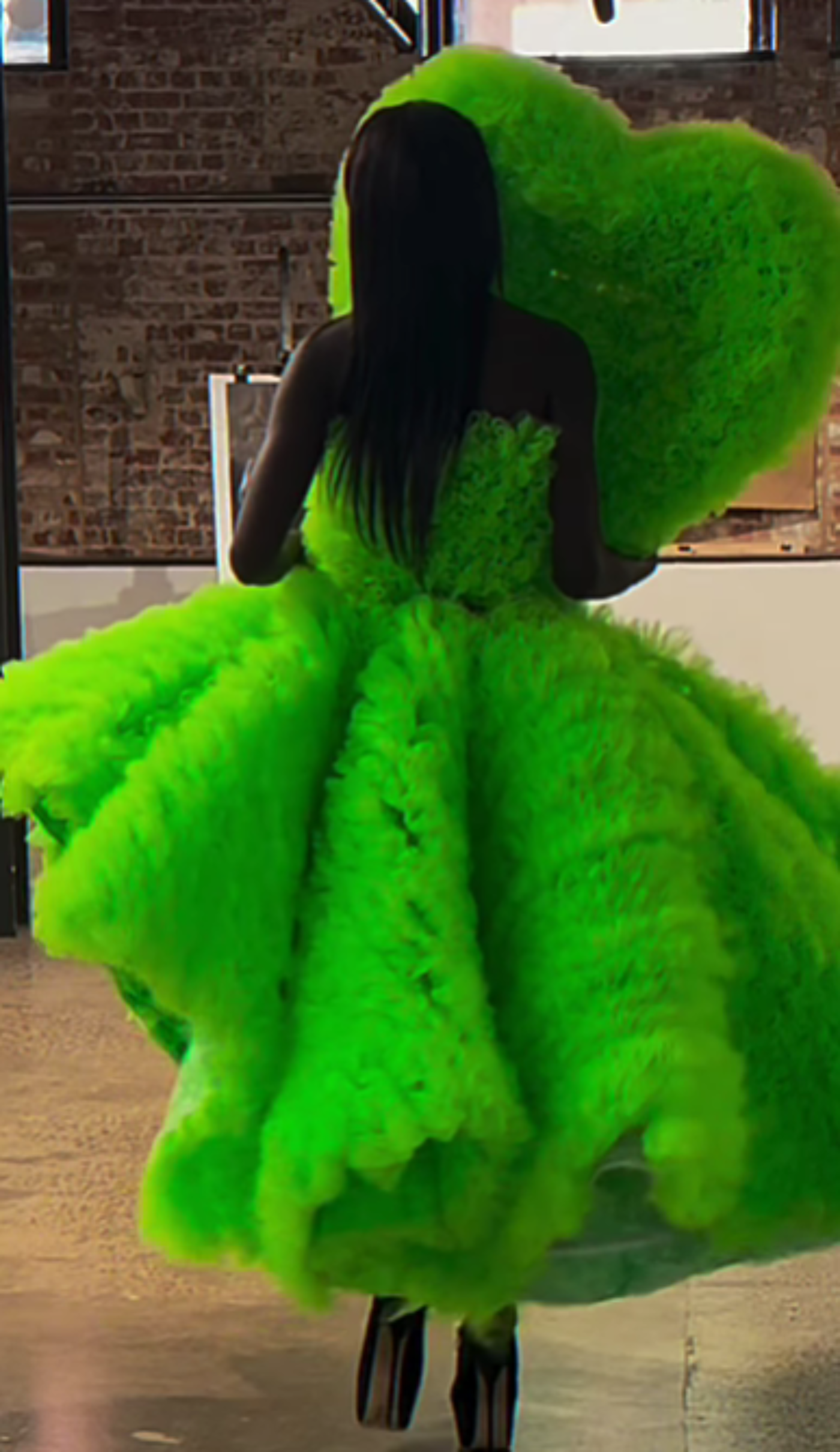 The image shows a person with dark skin and long black hair wearing a vibrant, fluffy, neon green dress. The dress has a large, heart-shaped detail on the back and a voluminous skirt, creating a striking and whimsical appearance.