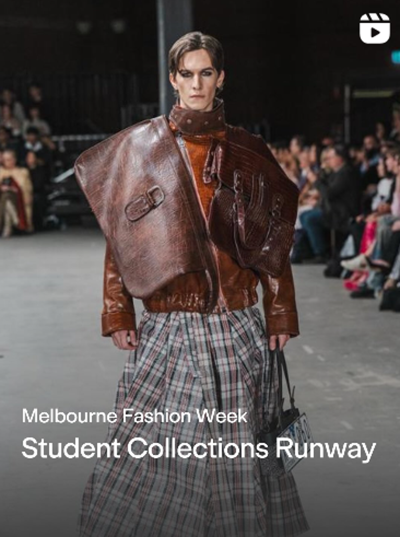 A person is walking on a runway wearing a brown jacket, a plaid skirt, and a large brown bag draped over their shoulder. The event is the Melbourne Fashion Week Student Collections Runway.