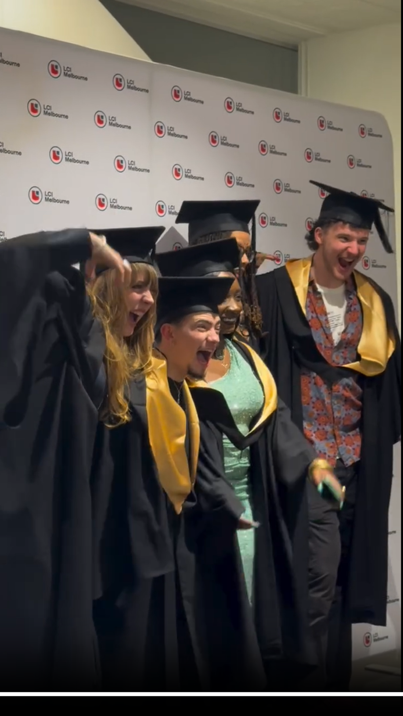A group of graduates in black gowns with gold accents pose for a photo in front of a white backdrop featuring a repeating logo.