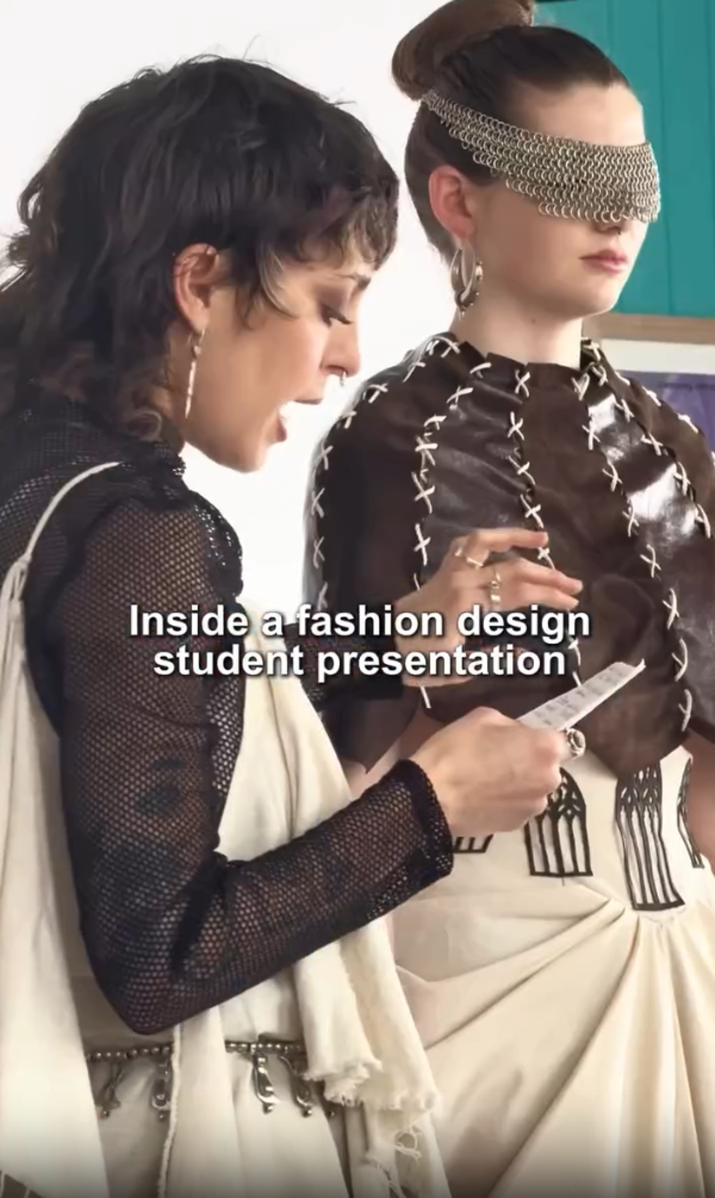 The image shows two fashion design students presenting their work. One student is holding a card and speaking, while the other is wearing a unique outfit with a chainmail-like blindfold. The presentation appears to be taking place indoors.