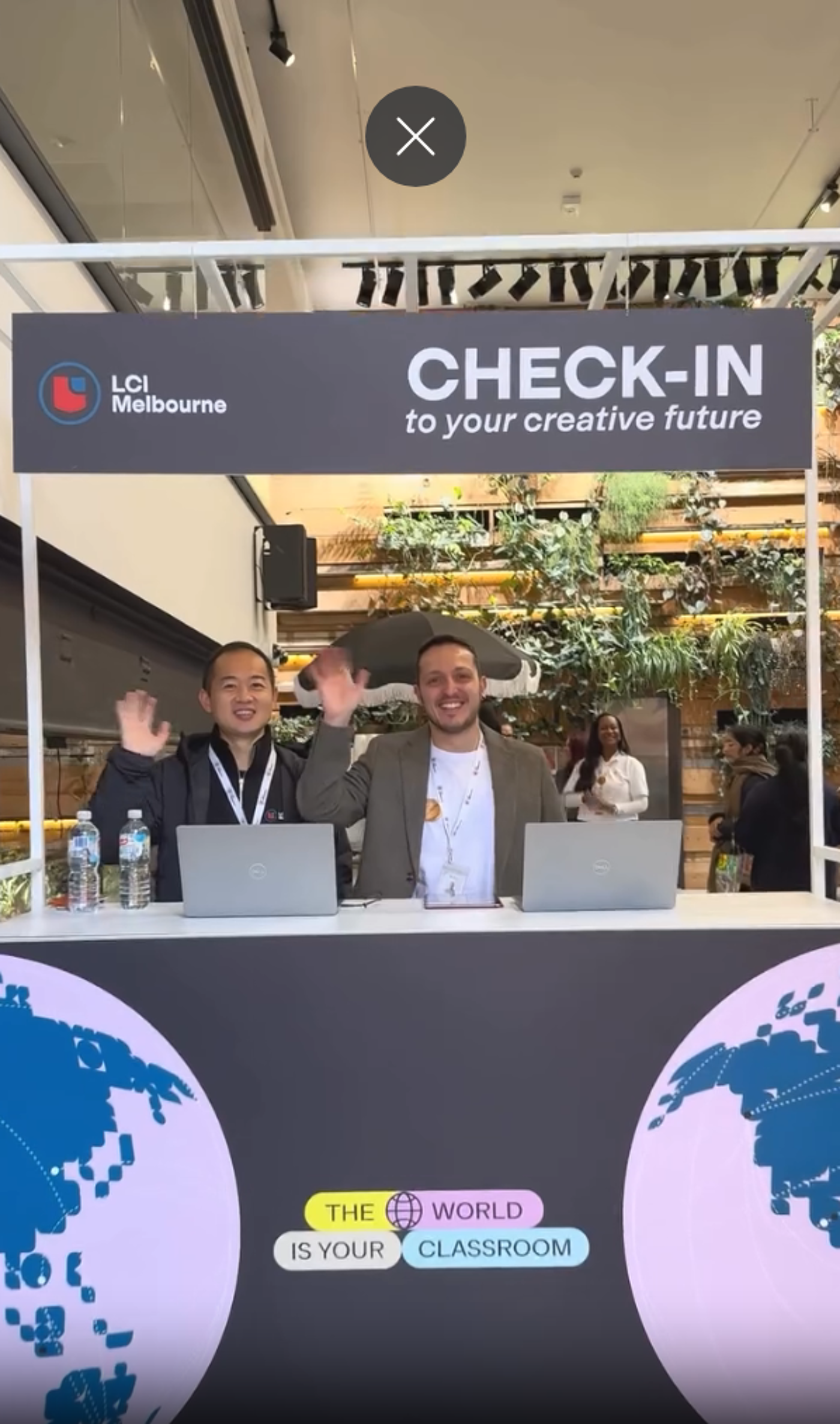 Two men are standing behind a check-in desk for LCI Melbourne, waving at the camera. The backdrop features the text "CHECK-IN to your creative future" and "THE WORLD IS YOUR CLASSROOM" with a globe graphic.