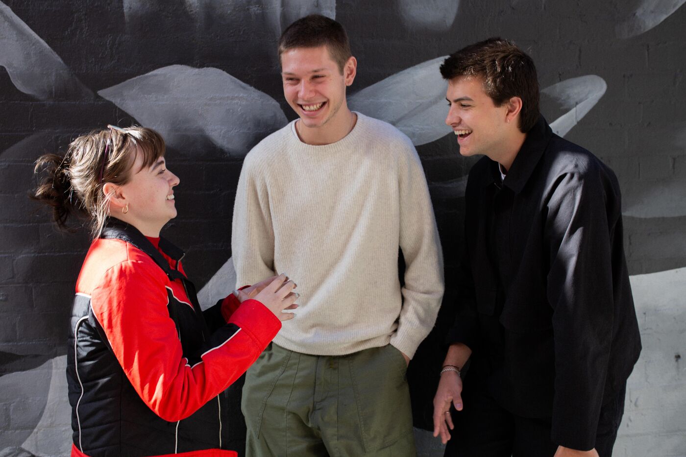 Three young adults standing together, laughing and engaging in a lively conversation. One wears a red jacket, another a beige sweater, and the third a black coat, with a dark mural wall in the background. The atmosphere is casual and cheerful.