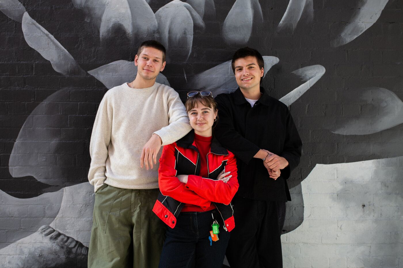 Three young artists pose confidently in front of a striking black and white mural, embodying the spirit of collaboration and youth.