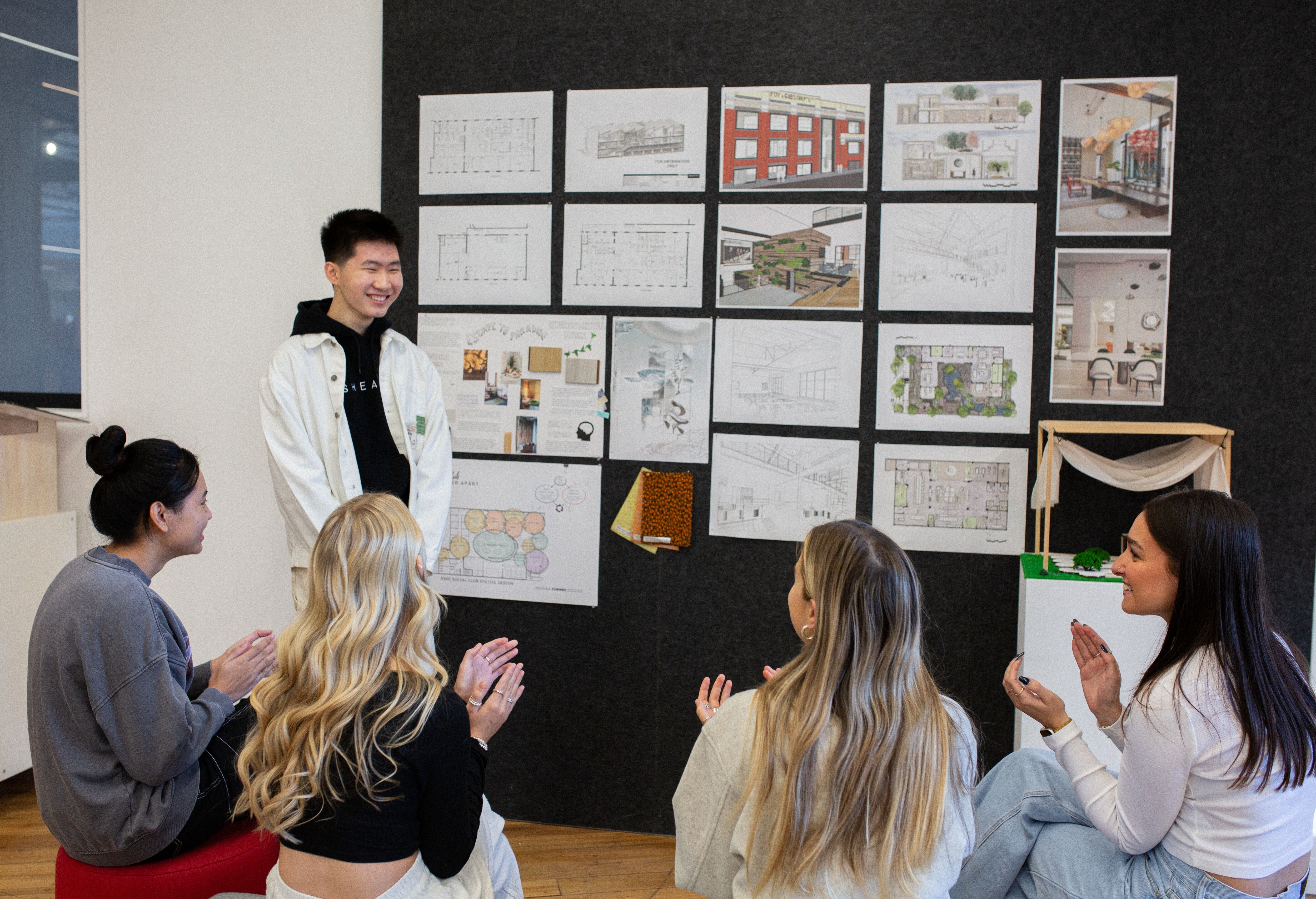 Un estudiante presenta diseños arquitectónicos a un grupo atento, exhibiendo planos detallados y modelos en una pared de galería.
