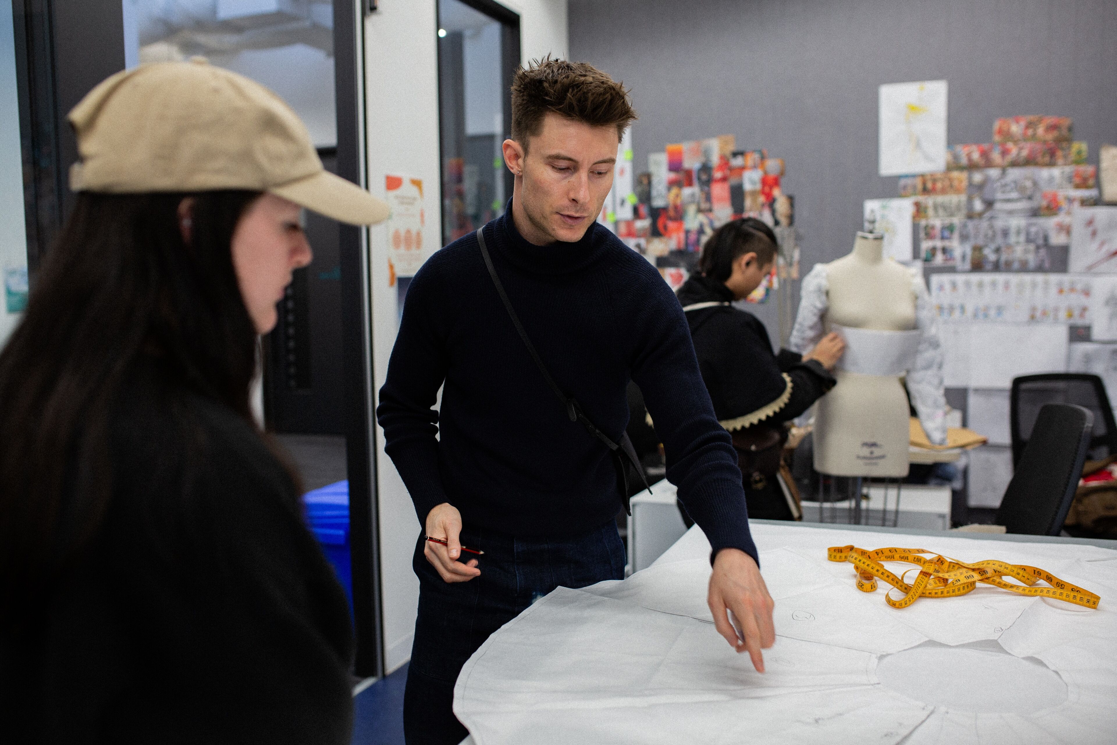 A focused fashion designer at work, detailing patterns on fabric with a mannequin and design sketches in the background.