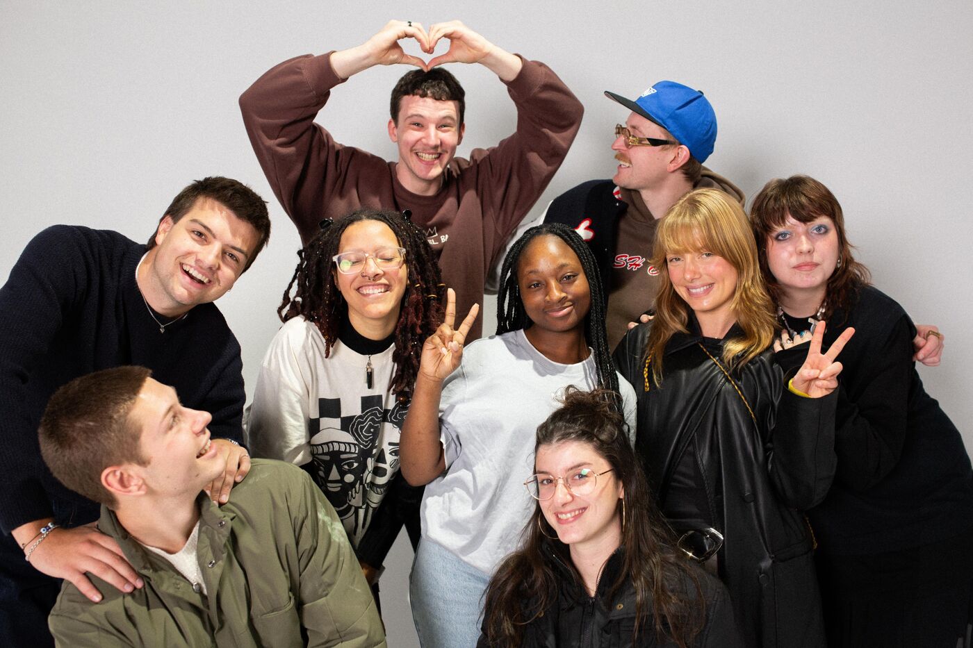 A diverse group of young people posing for a cheerful photo. They express joy and camaraderie, with some making heart and peace signs. The bright smiles and casual clothing create a lively and warm atmosphere against a neutral background.