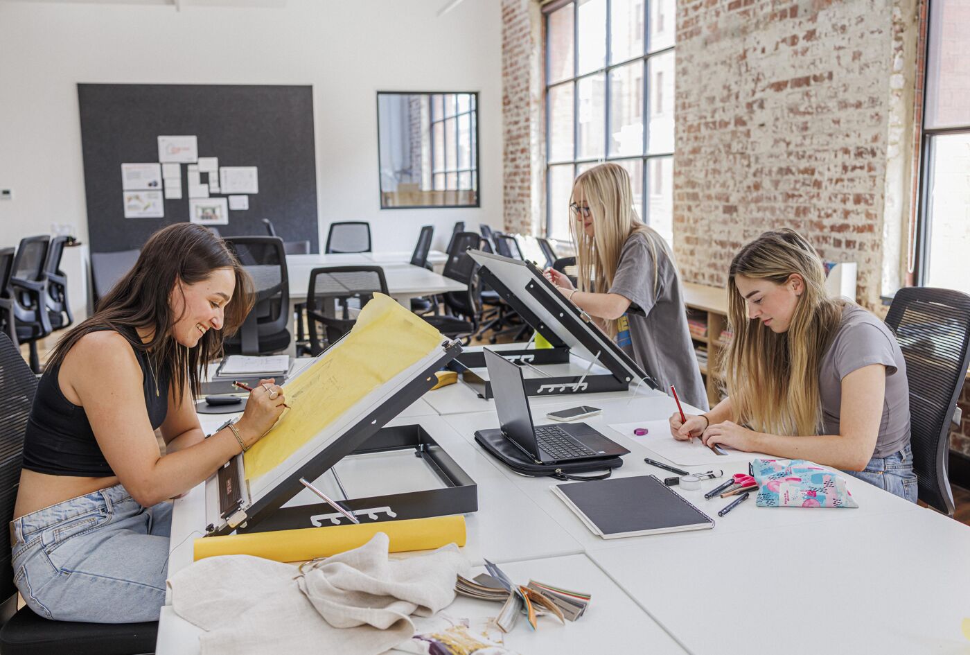 Three young women engaged in creative work in a modern design studio. They are using drafting tables to sketch, surrounded by tools and laptops, with natural light streaming through large windows. The atmosphere is focused and collaborative.